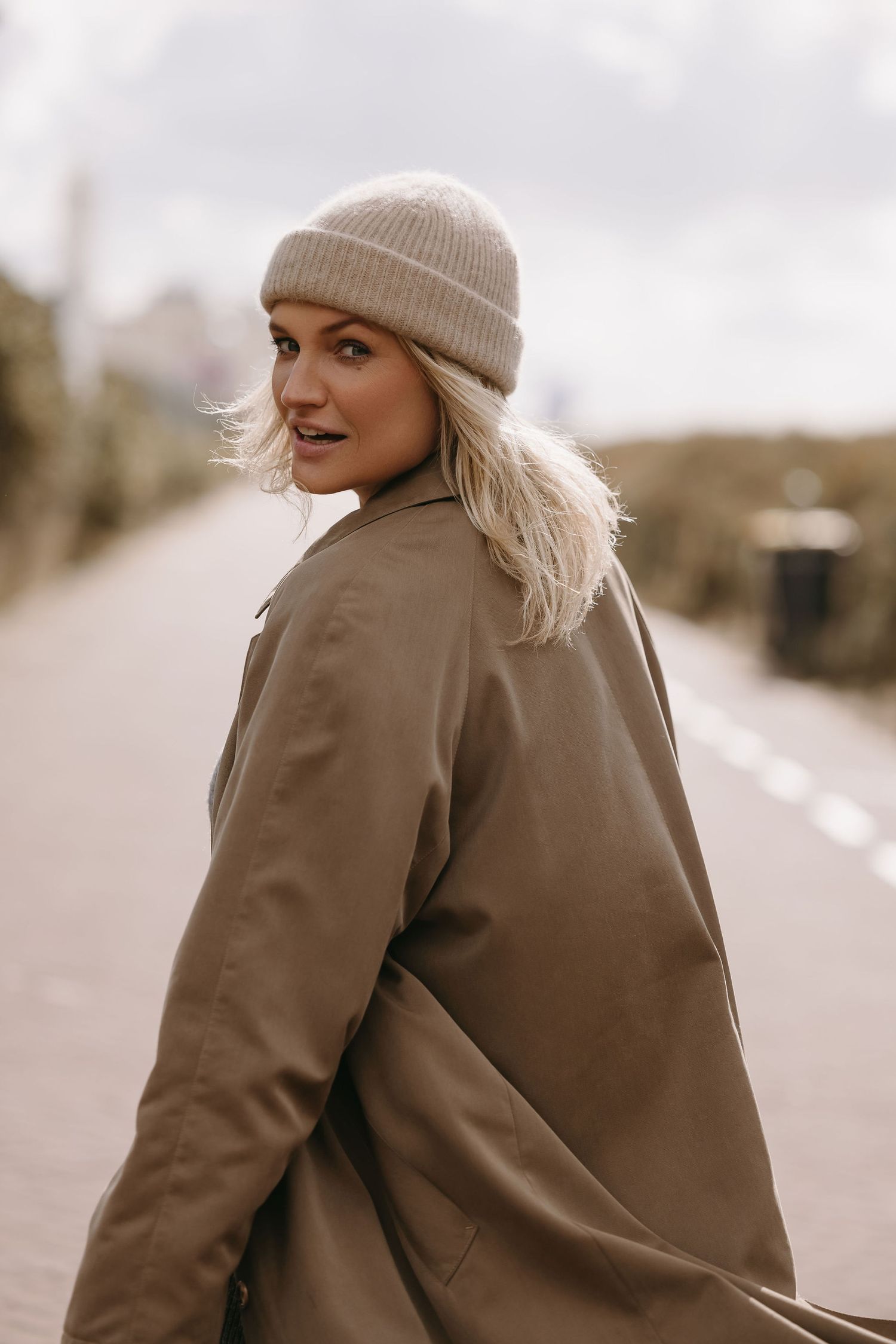 Stylish autumn portrait of a person in a beige beanie and brown coat looking over their shoulder on a misty road.