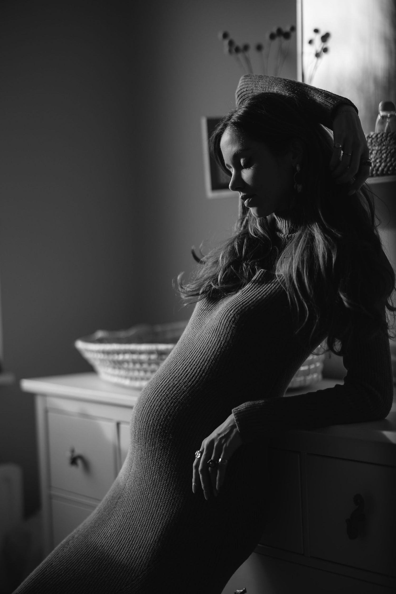 Black and white portrait of a pregnant woman sitting in silhouette by a window.