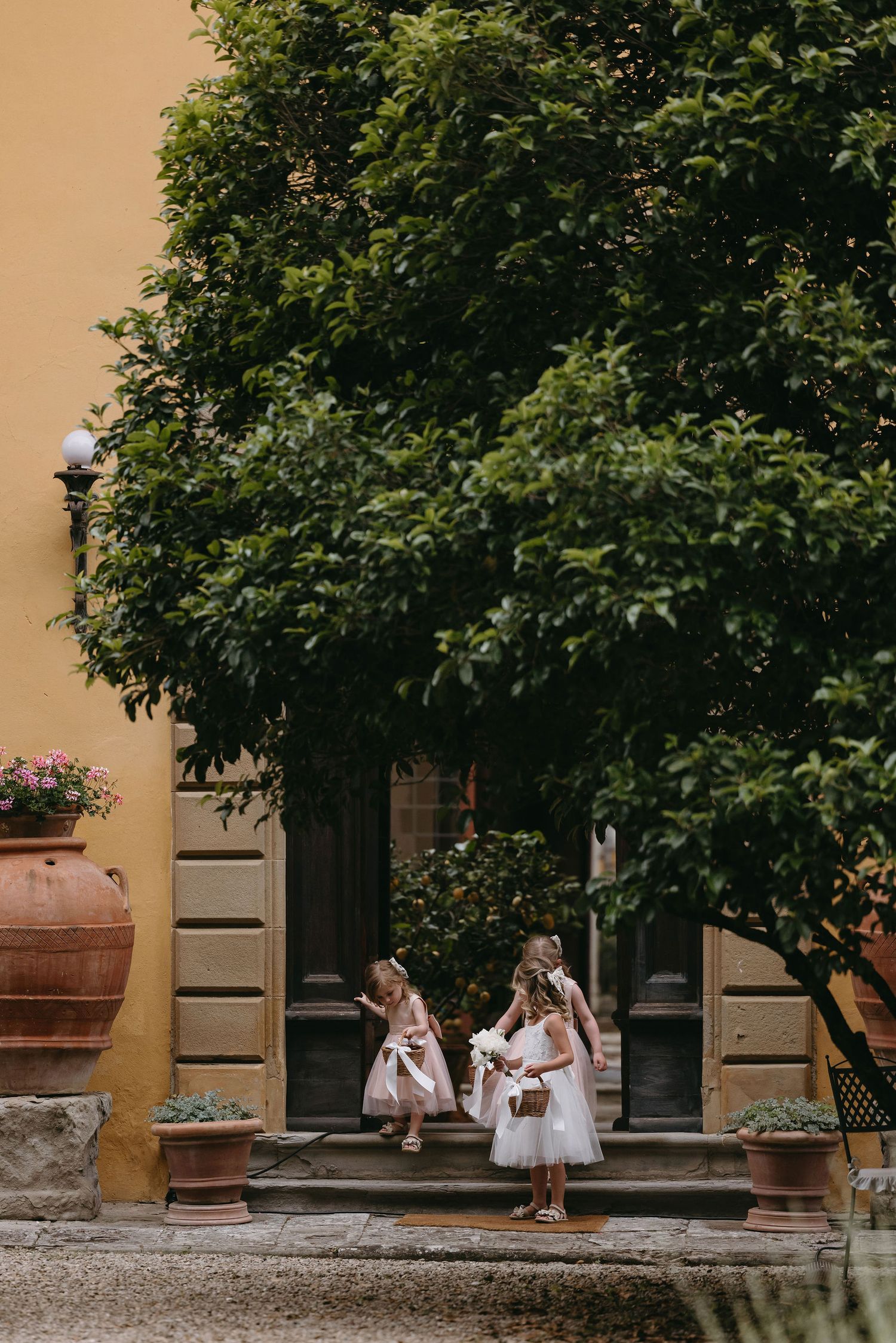 Young attendants in white dresses standing outside an Italian villa with terracotta planters.