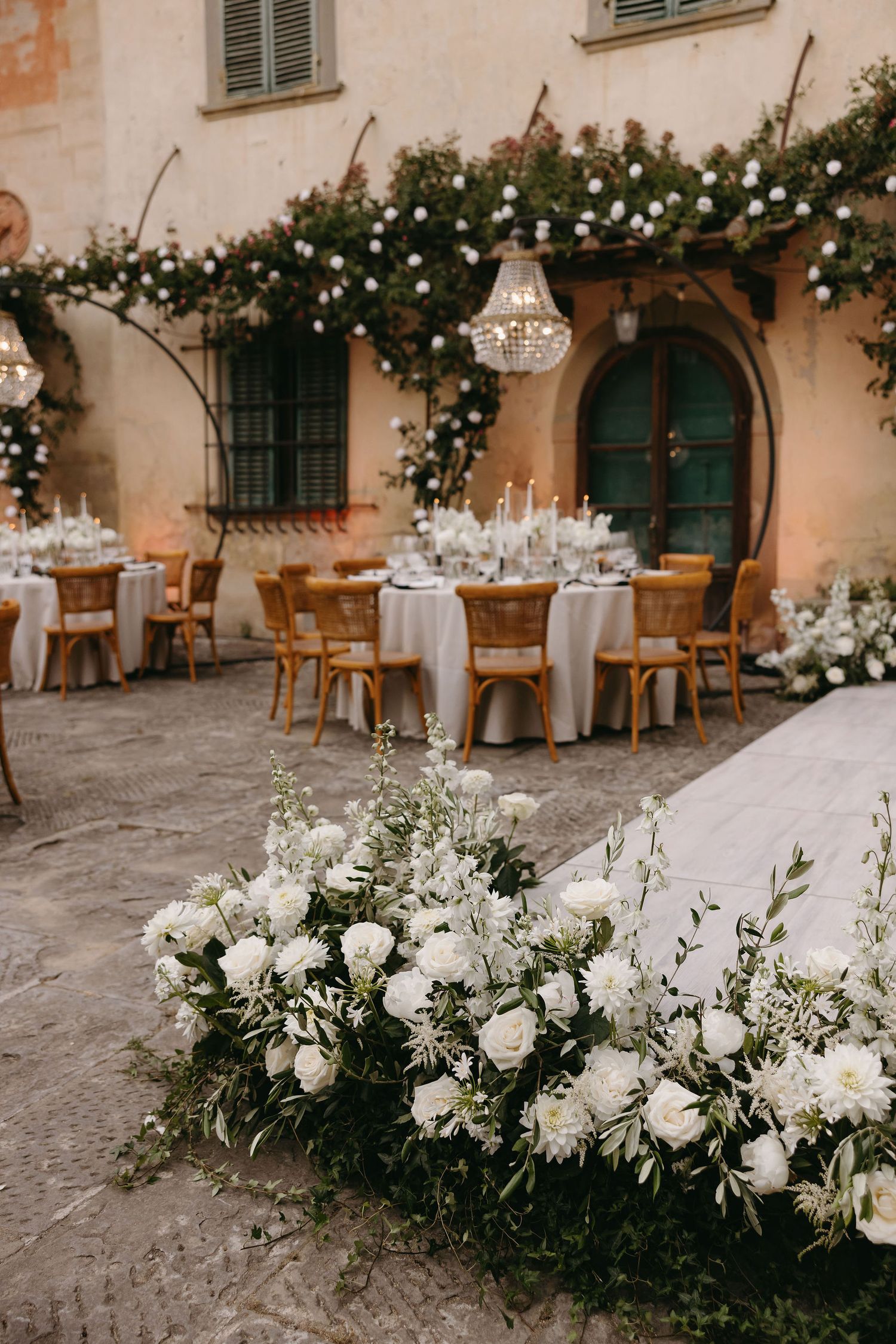 Romantic outdoor wedding reception setup with white flowers, crystal chandeliers and wooden chairs.