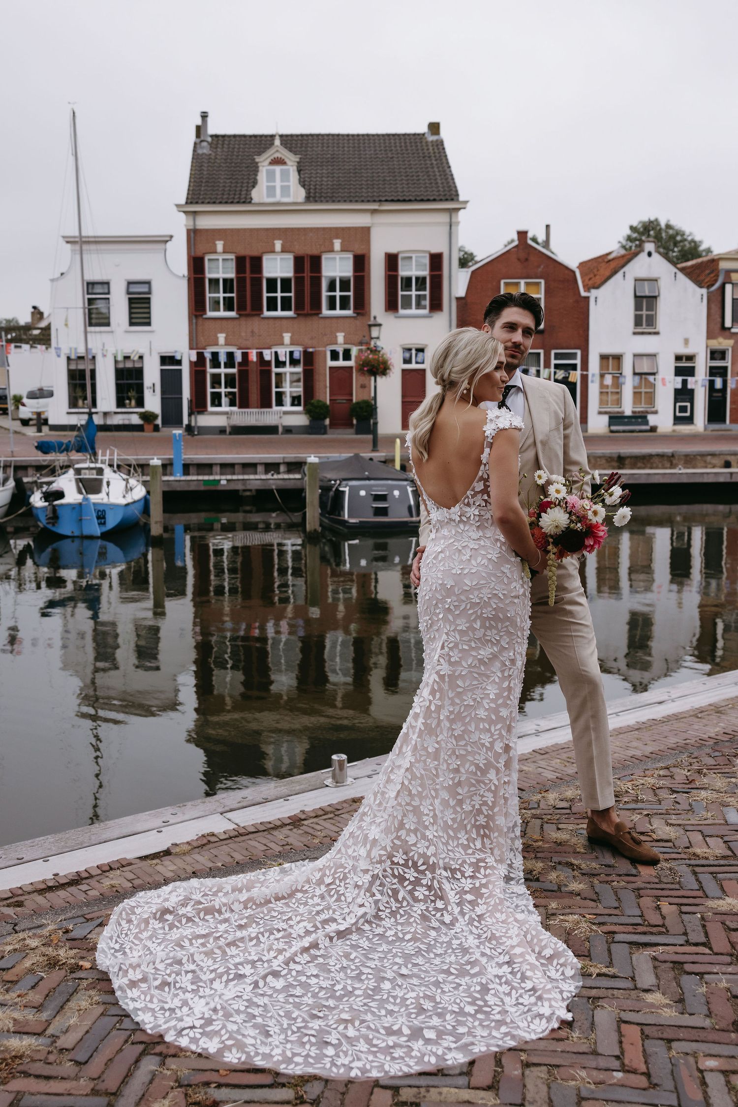Wedding dress with intricate lace train photographed against historic waterfront buildings.