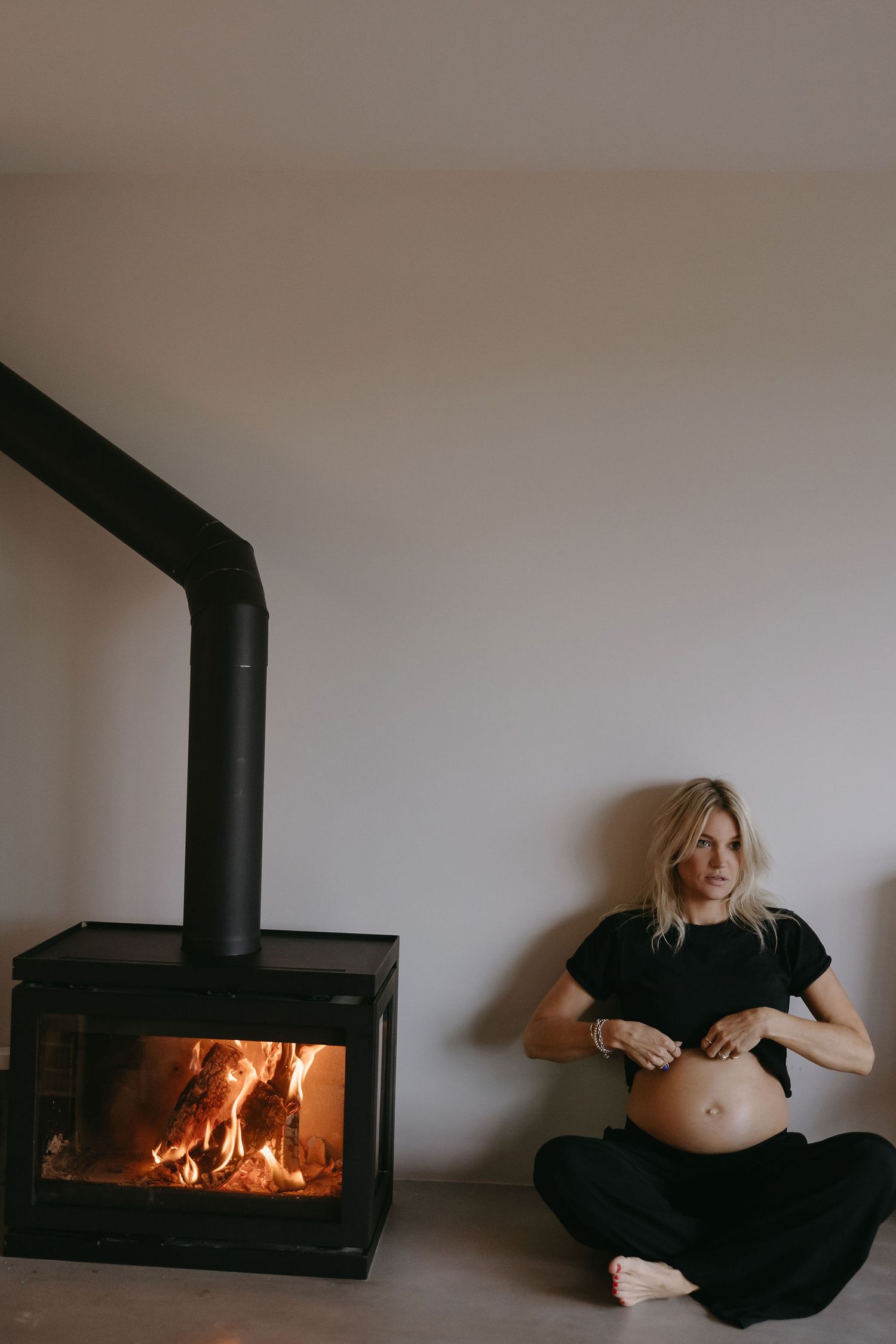 Cozy maternity photo by wood stove showing pregnant belly in black outfit.