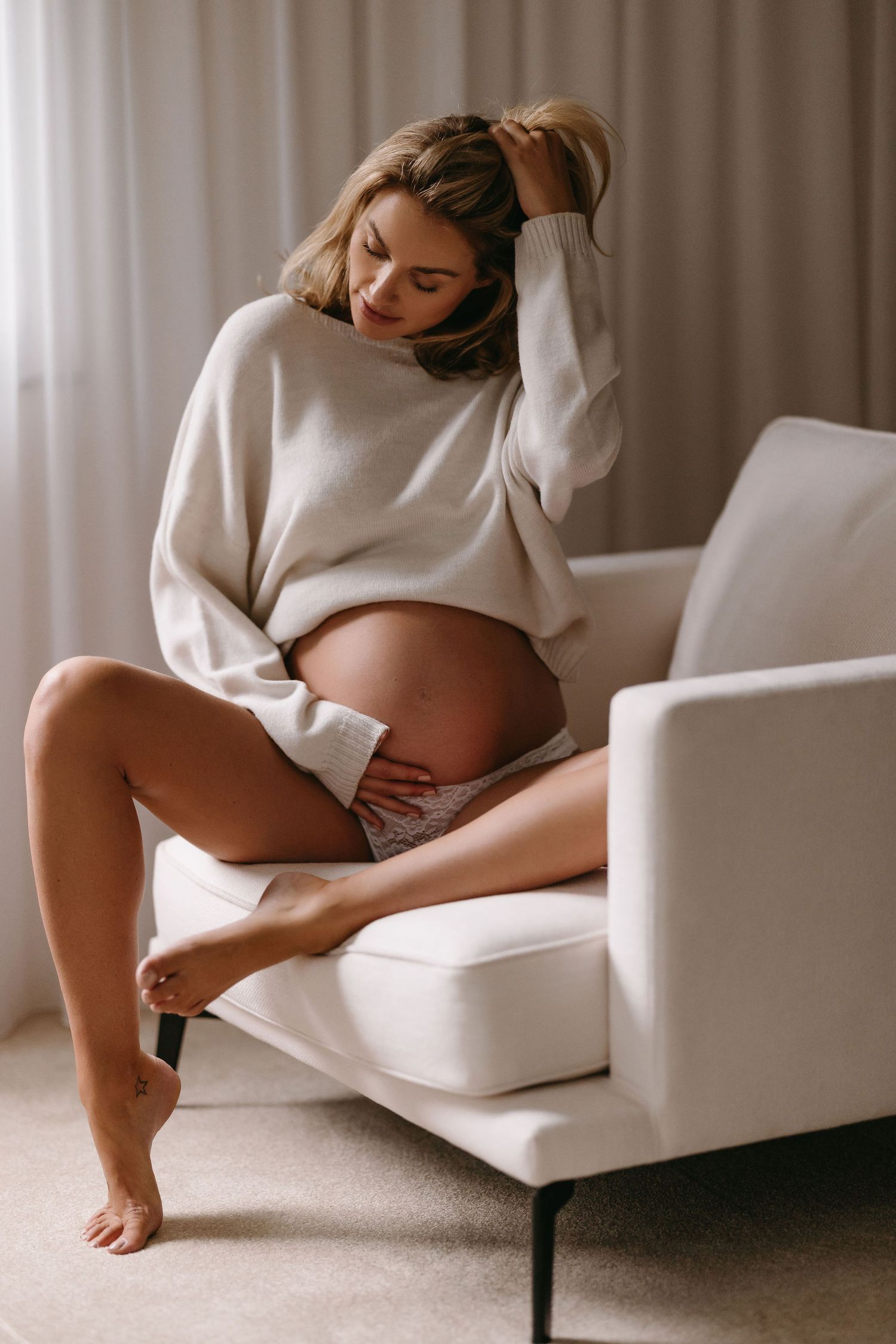 Woman in cream sweater lounging on white armchair during pregnancy photoshoot.
