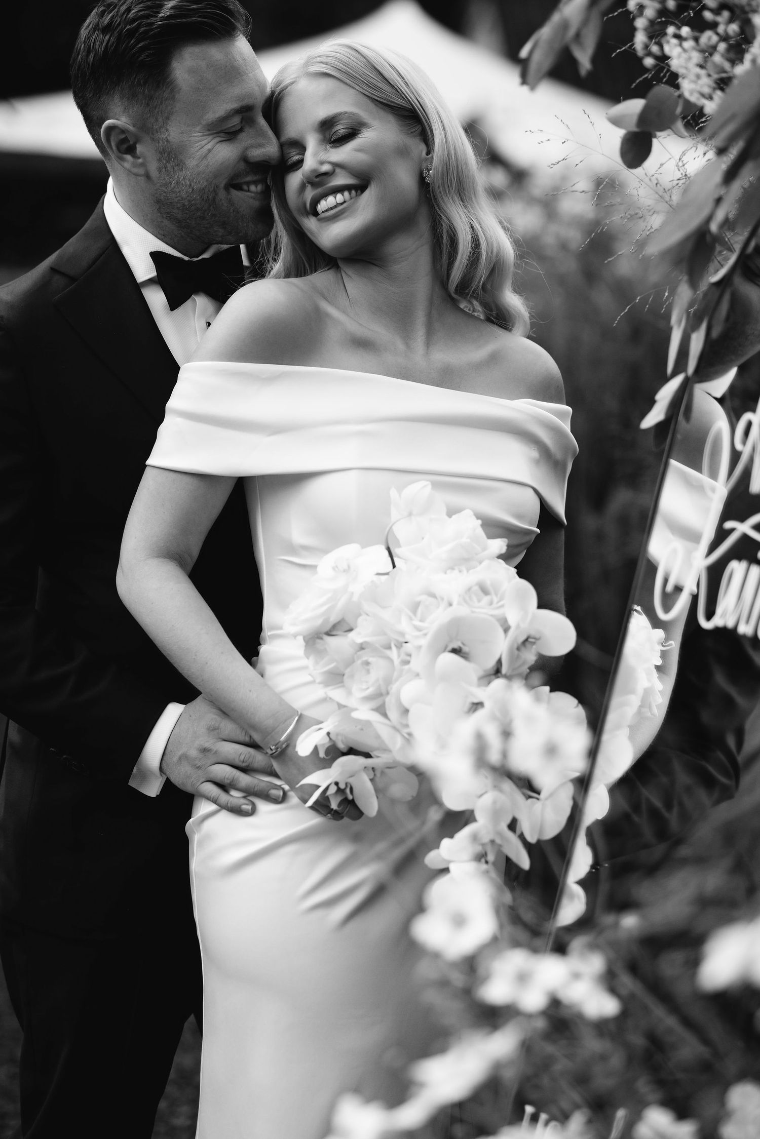 Intimate black and white portrait of a couple embracing in elegant wedding attire with white floral bouquet.