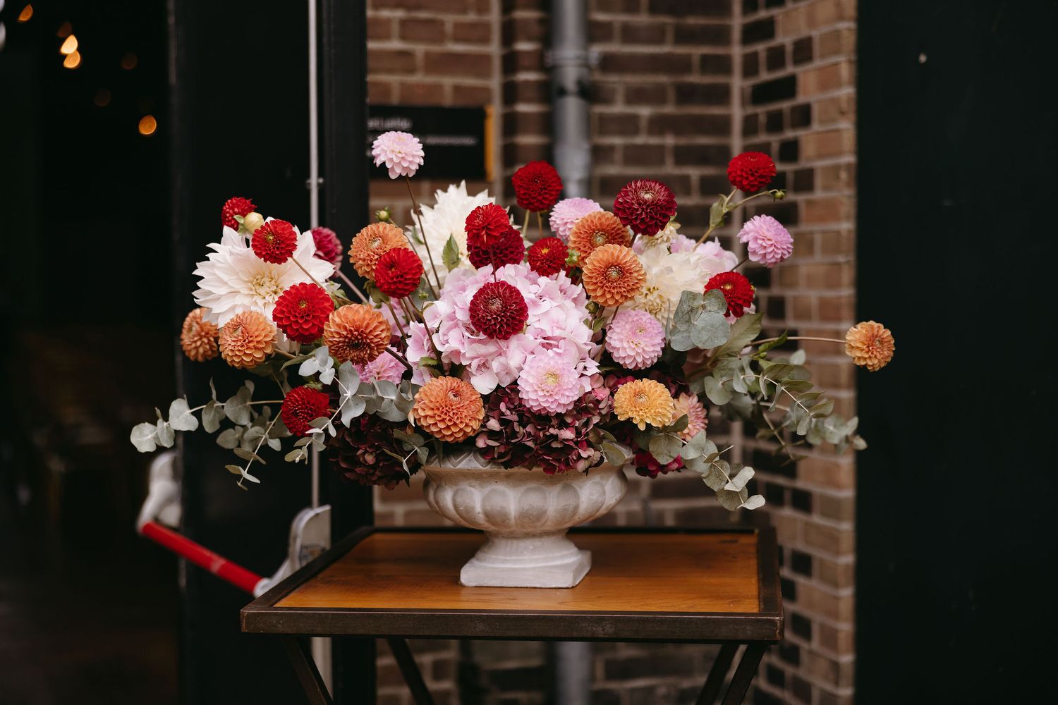 Elegant floral centerpiece with red and pink blooms in a white vase against exposed brick wall.