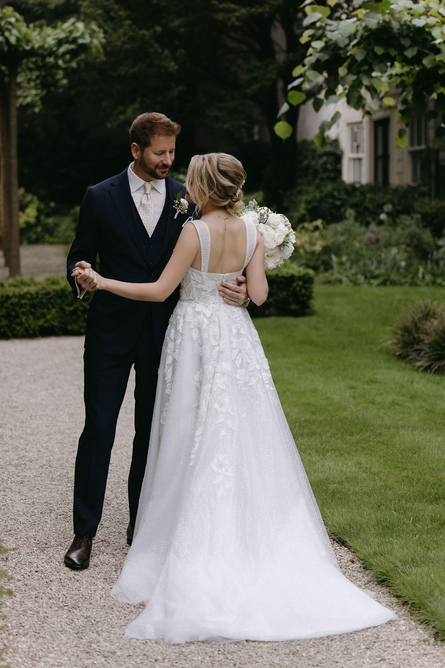 A romantic garden wedding photo showing the back of a bride and groom in formal attire on a gravel path.