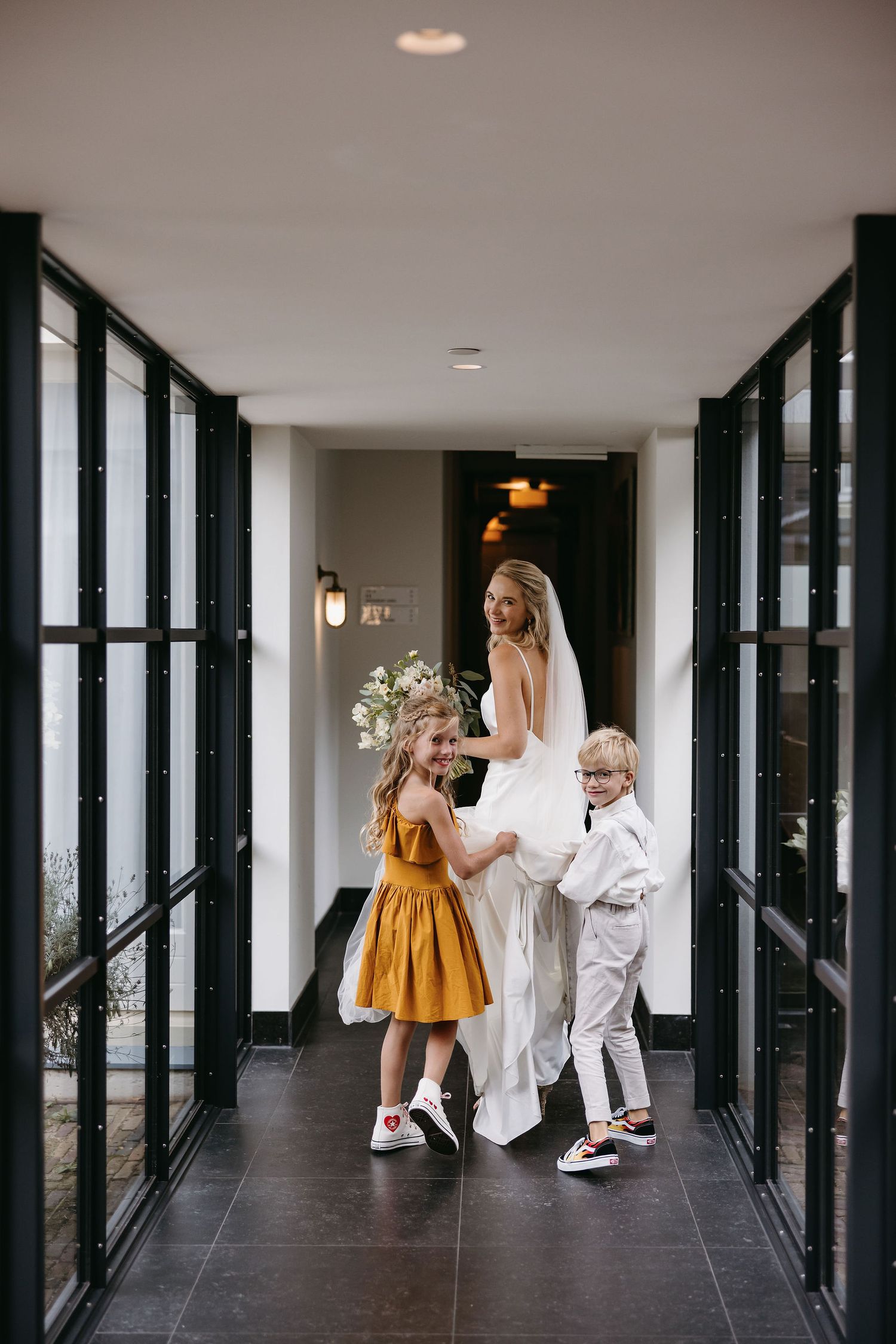 Bride in white dress walks hallway with children in formal attire.