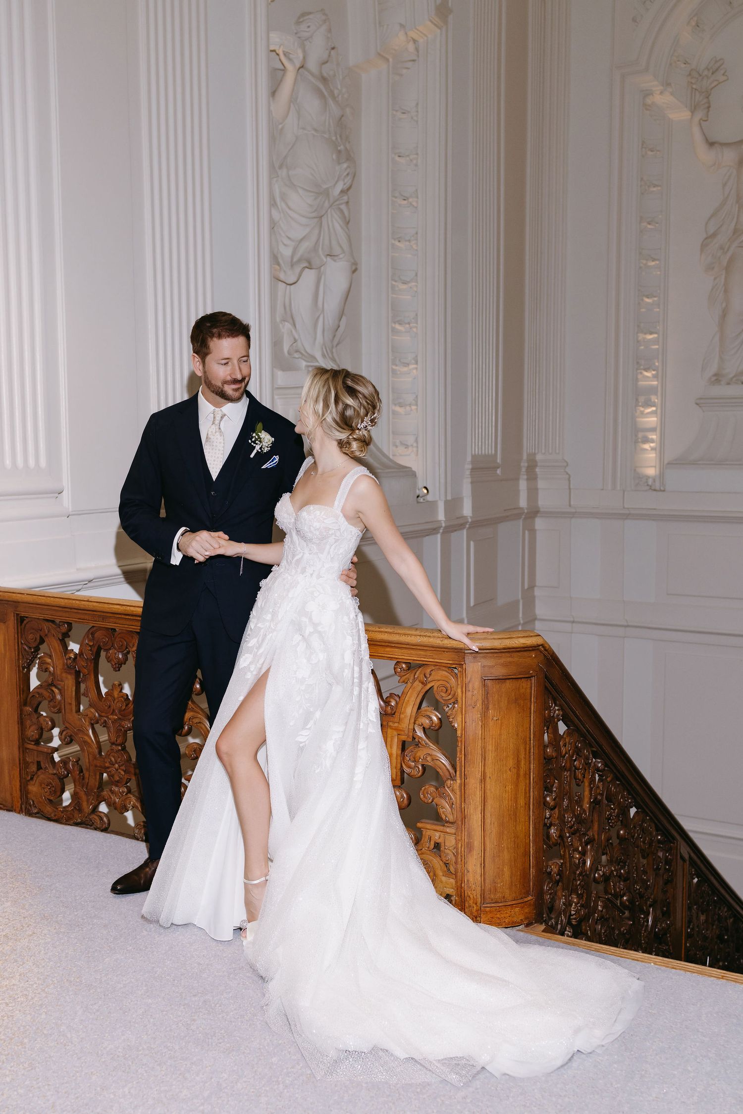 A couple in wedding attire dances together on a grand marble and wood staircase in an elegant setting.