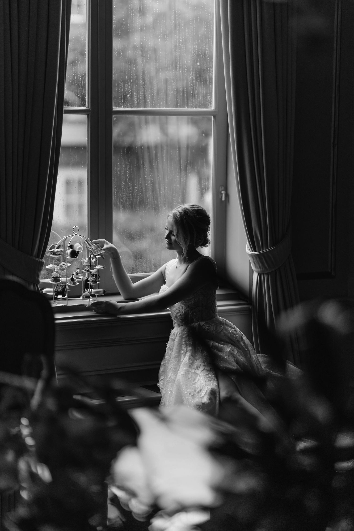 A silhouetted figure in a flowy dress sits beside a rainy window with dramatic curtains in this black and white photograph.
