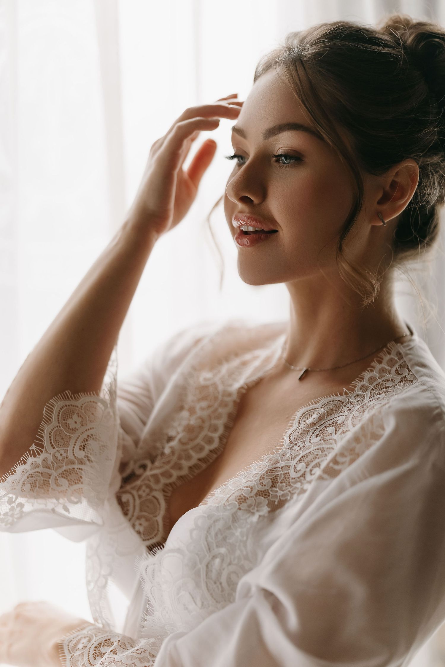 A romantic portrait of a bride wearing a delicate white lace robe while getting ready by a window in soft lighting.