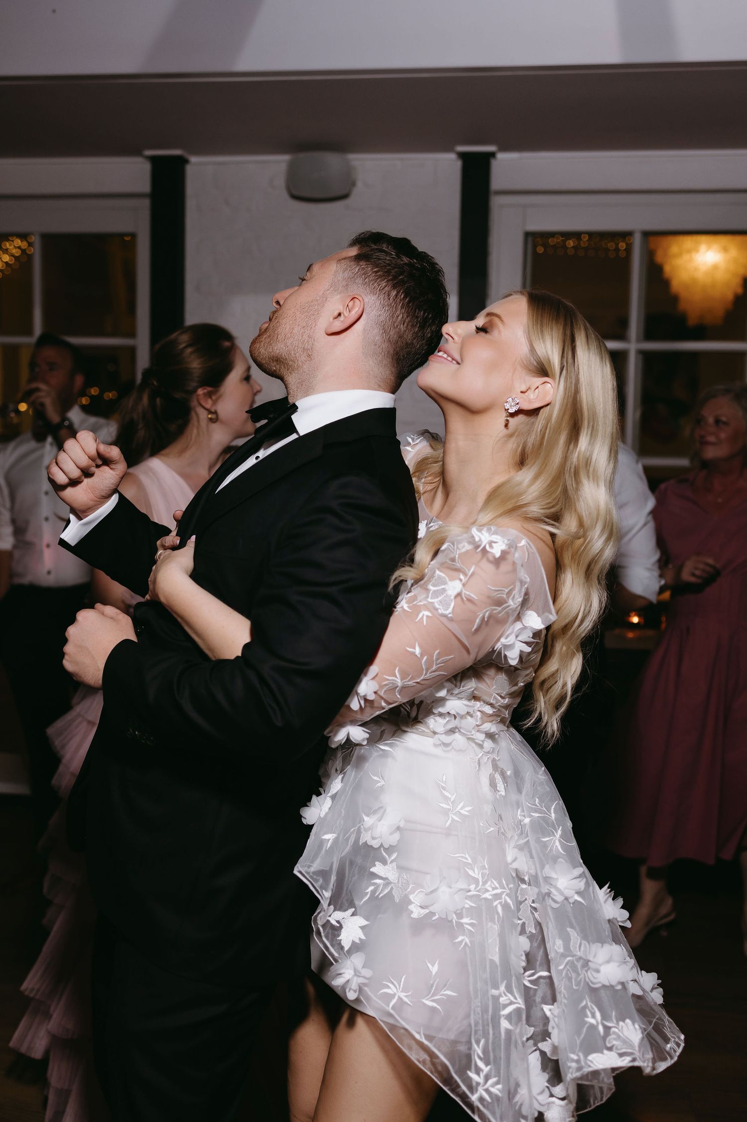 Couple dancing enthusiastically at wedding reception, bride wearing delicate lace overlay dress.