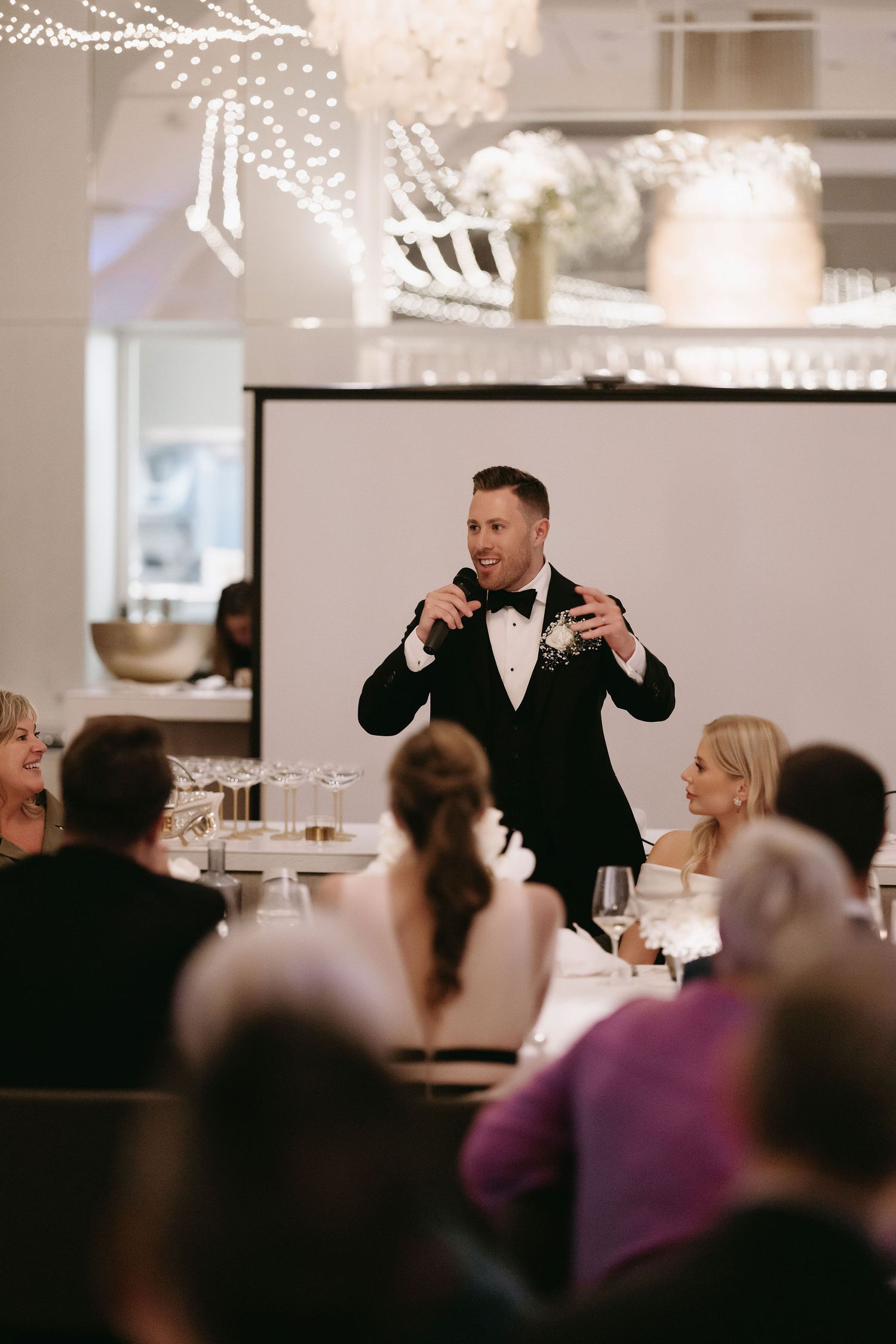 A speaker in a tuxedo addresses wedding guests during an indoor reception.