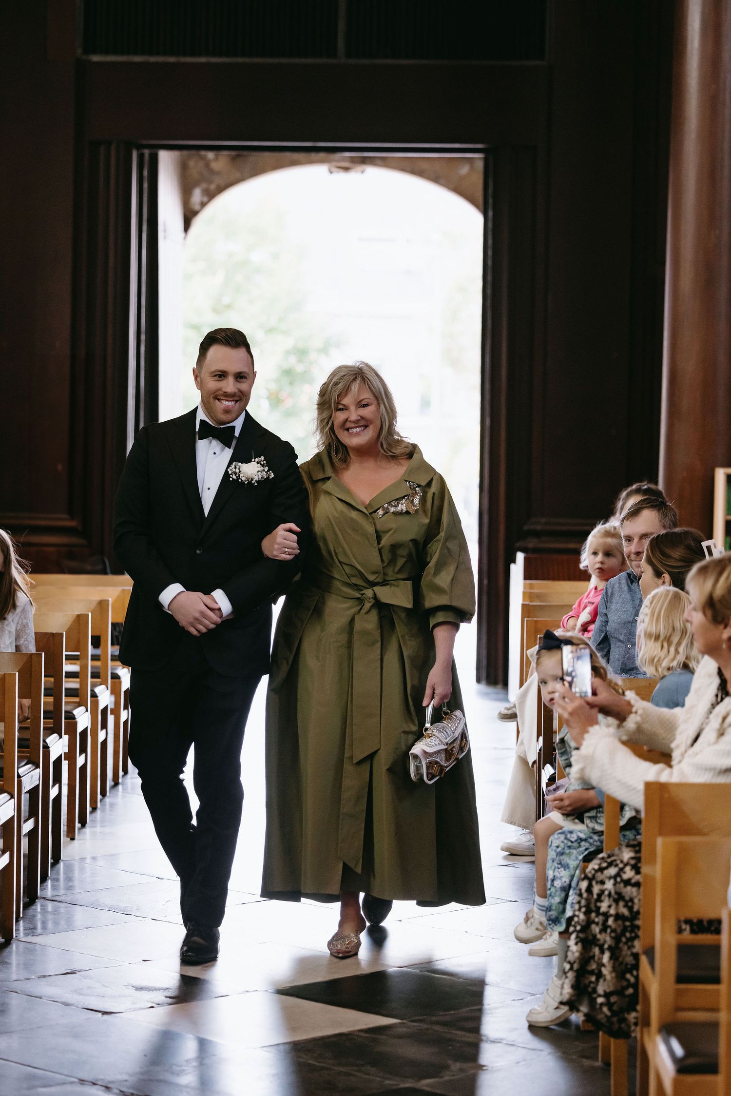 A church processional with guests seated in wooden pews.