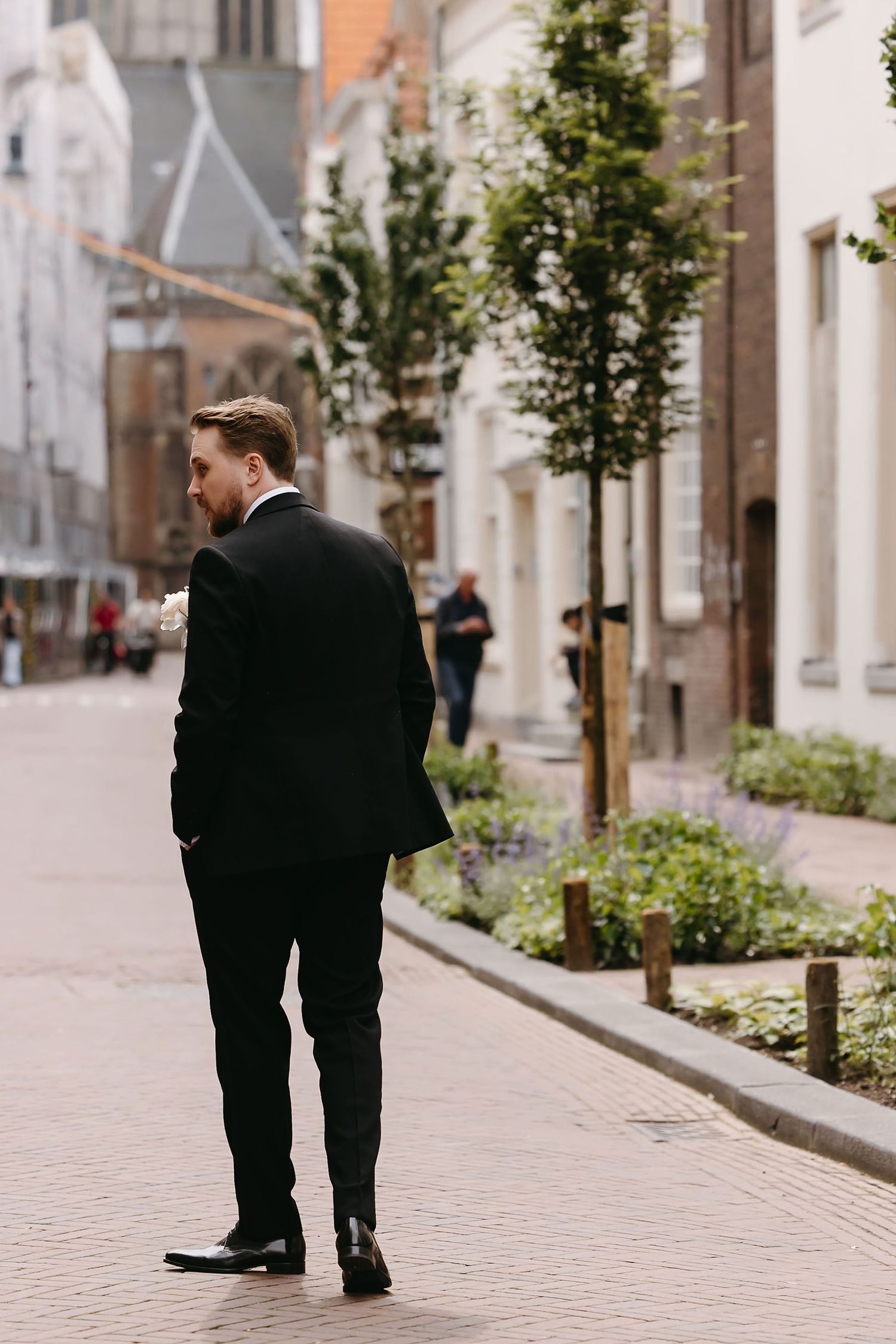 A person in a black suit stands on a brick-paved street lined with historic buildings and young trees.