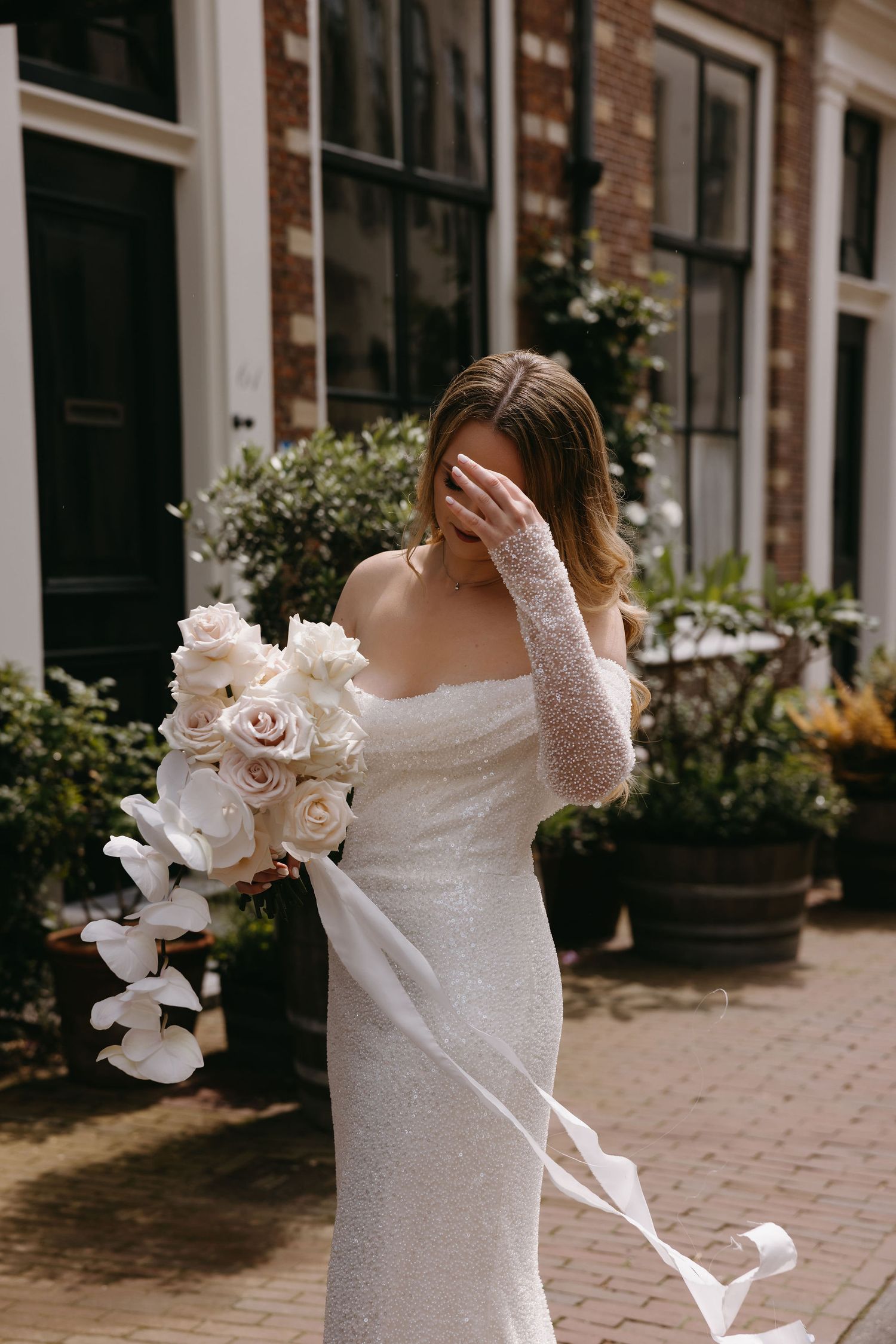 A bride in a fitted white dress holds a cascading bouquet of white orchids on a brick-lined street.