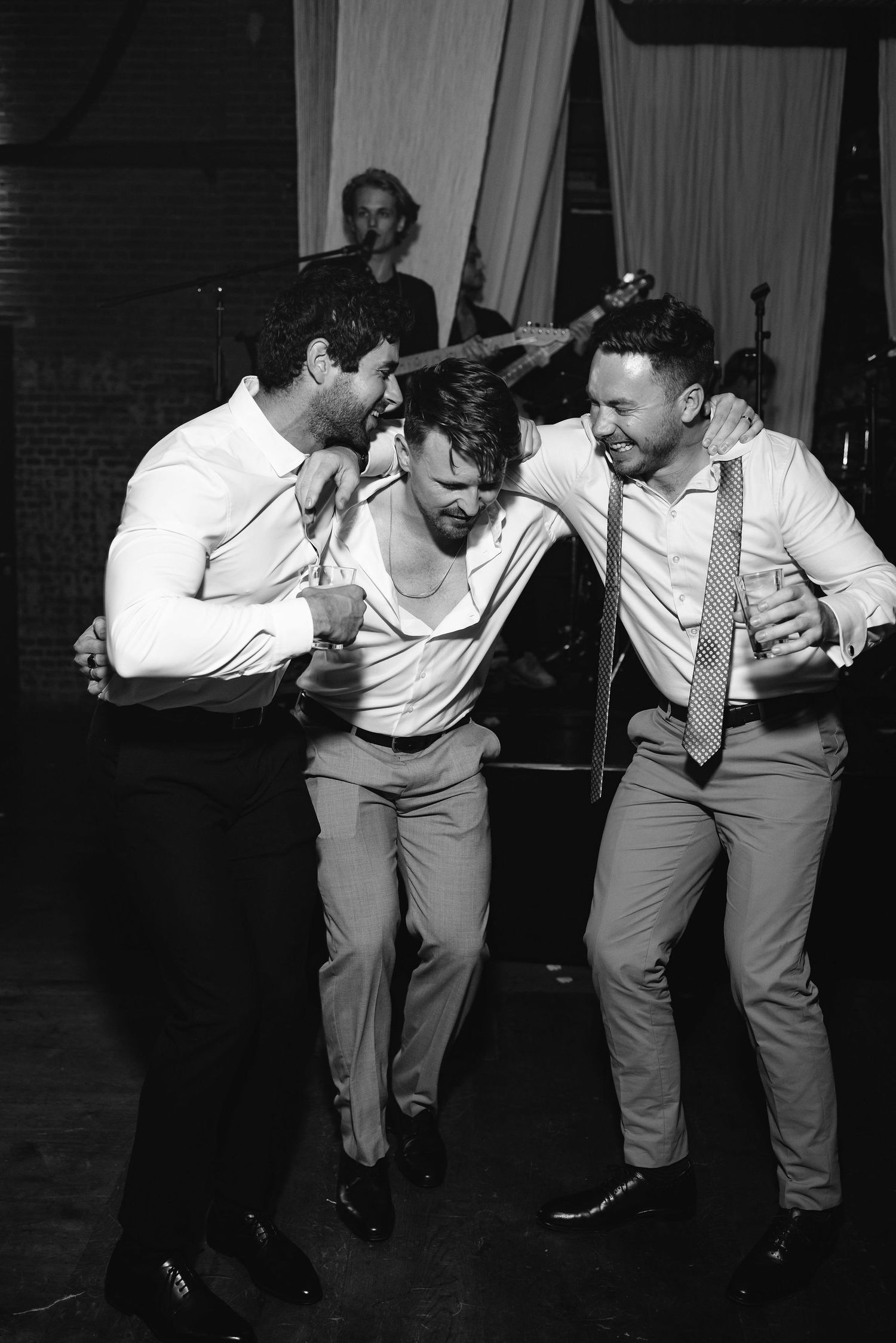 Black and white photo of three friends dancing and laughing together at a wedding reception.