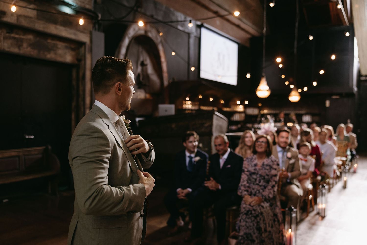Guest speaking to wedding attendees in an industrial-style venue with string lights overhead.