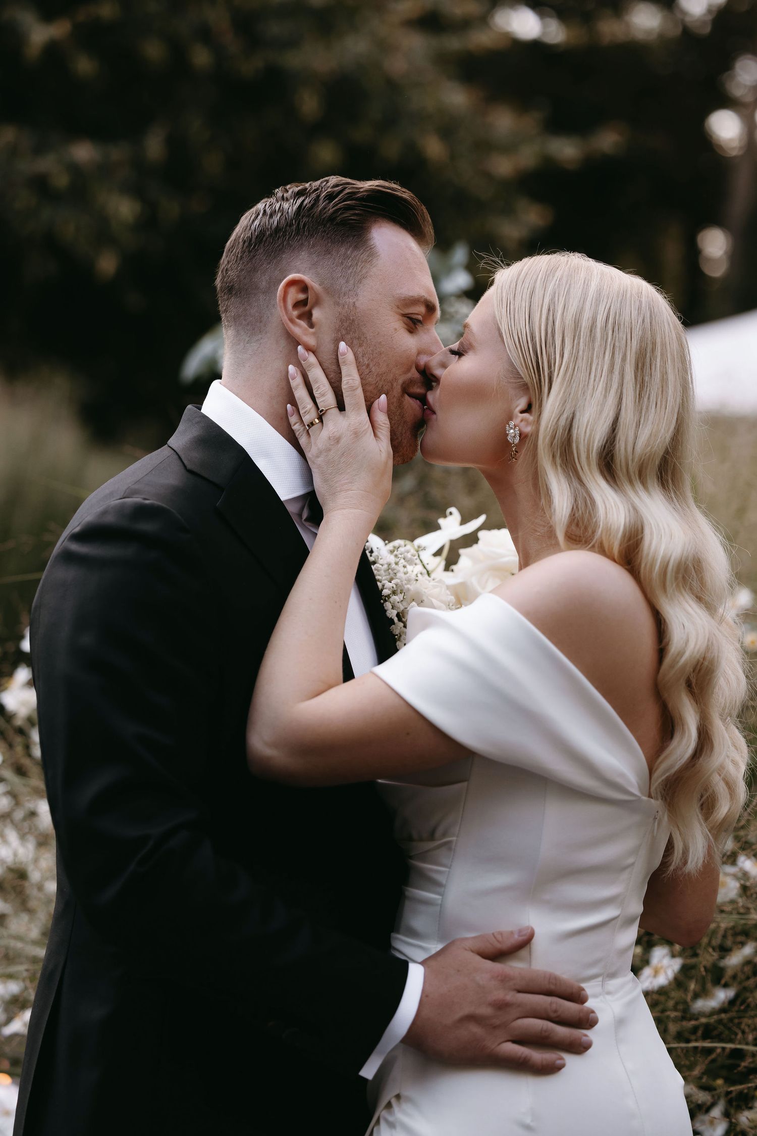 A romantic wedding kiss between newlyweds in elegant attire against a natural outdoor backdrop.