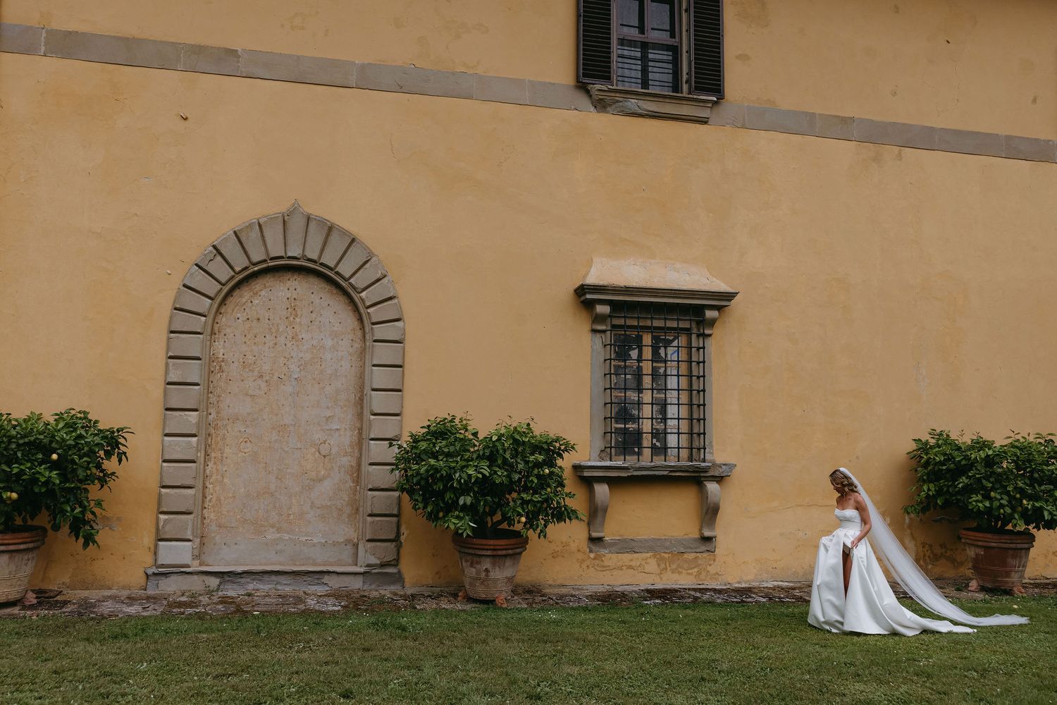 A white wedding dress flowing on the green grass beside a historic yellow building with arched doorway and potted plants.