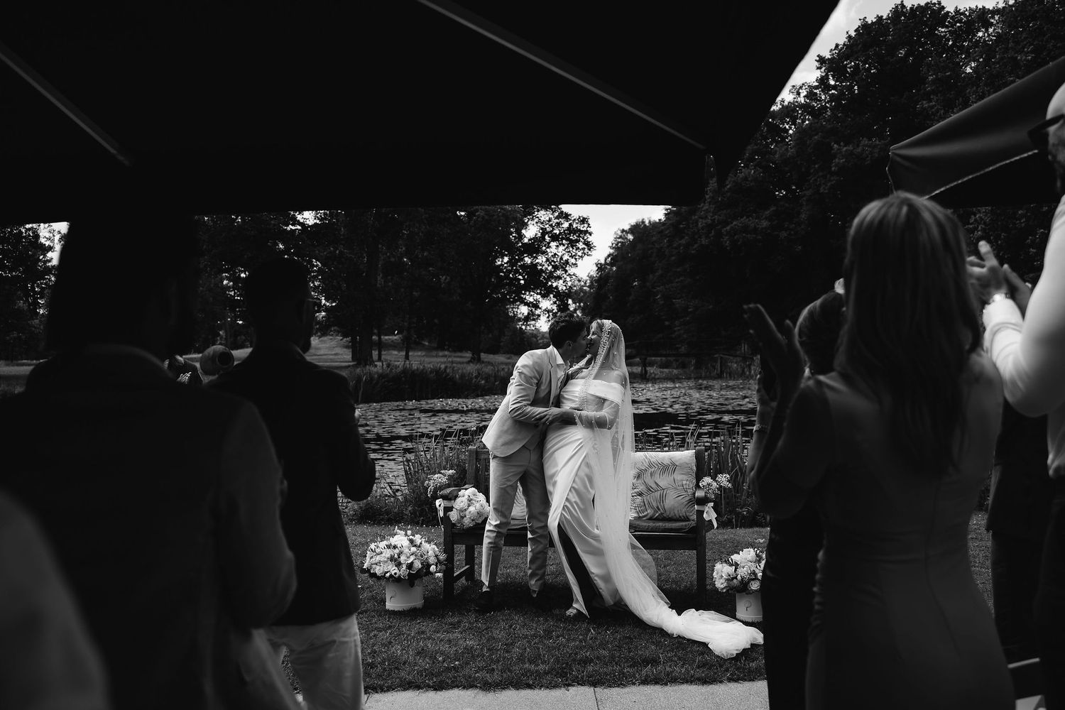 Black and white candid photo of a wedding couple during an outdoor ceremony with guests in silhouette.