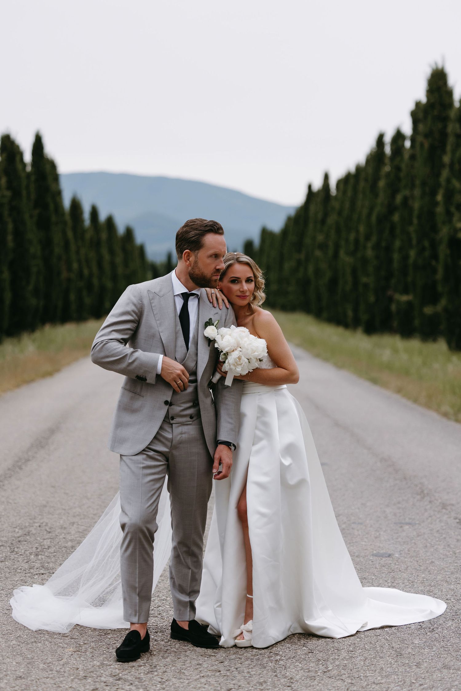 A romantic wedding photo series showing a couple in formal attire posing playfully on a remote tree-lined country road.