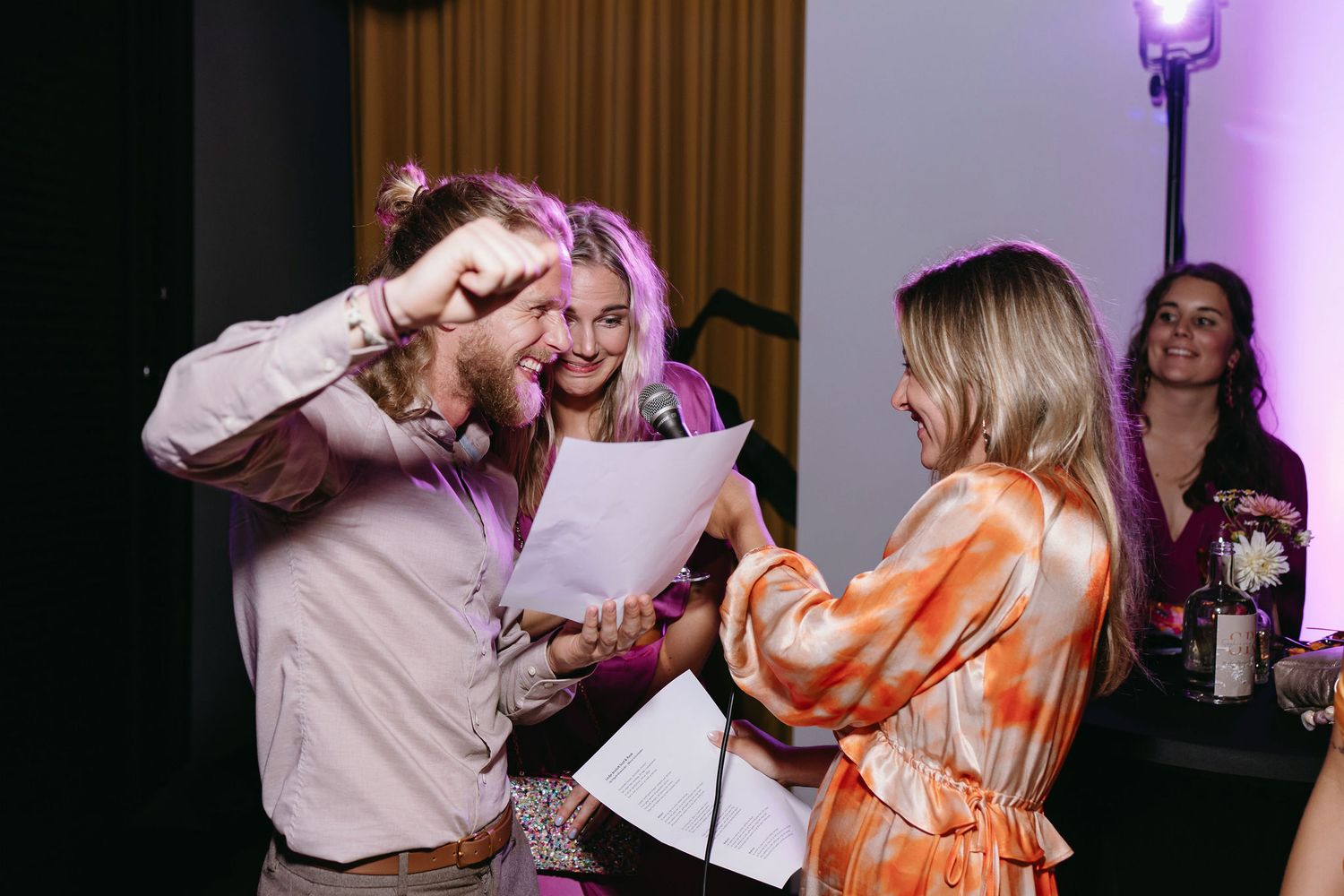 Wedding celebration with guests singing and reading from papers in purple-lit indoor setting.