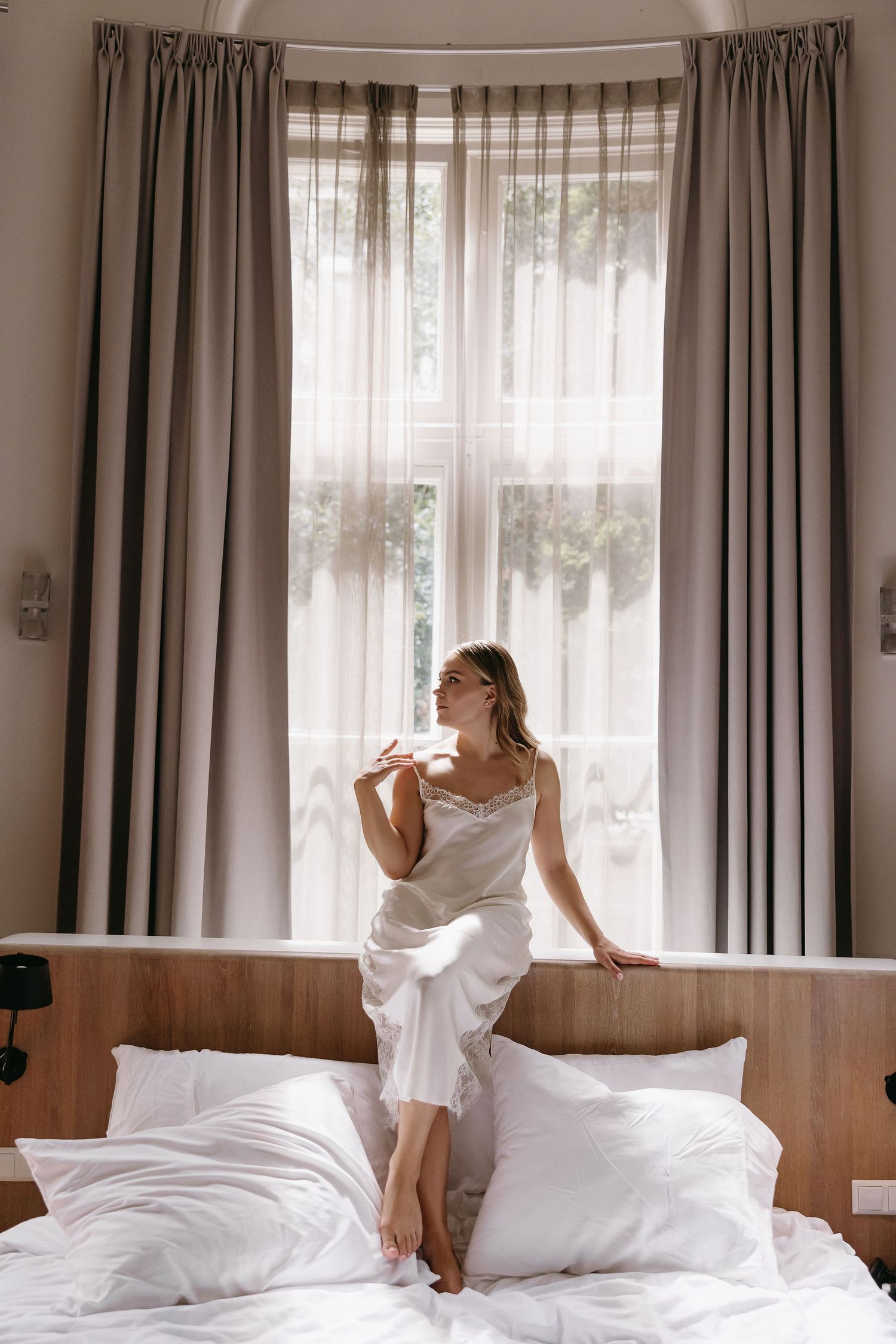 Sunlit scene of someone in white seated on a bed by a window with flowing curtains.