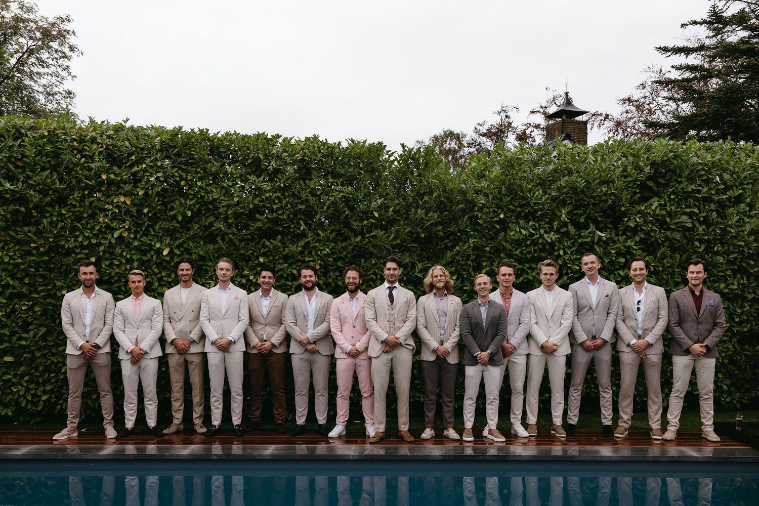 Large wedding party in light colored suits lined up by swimming pool for group photo.