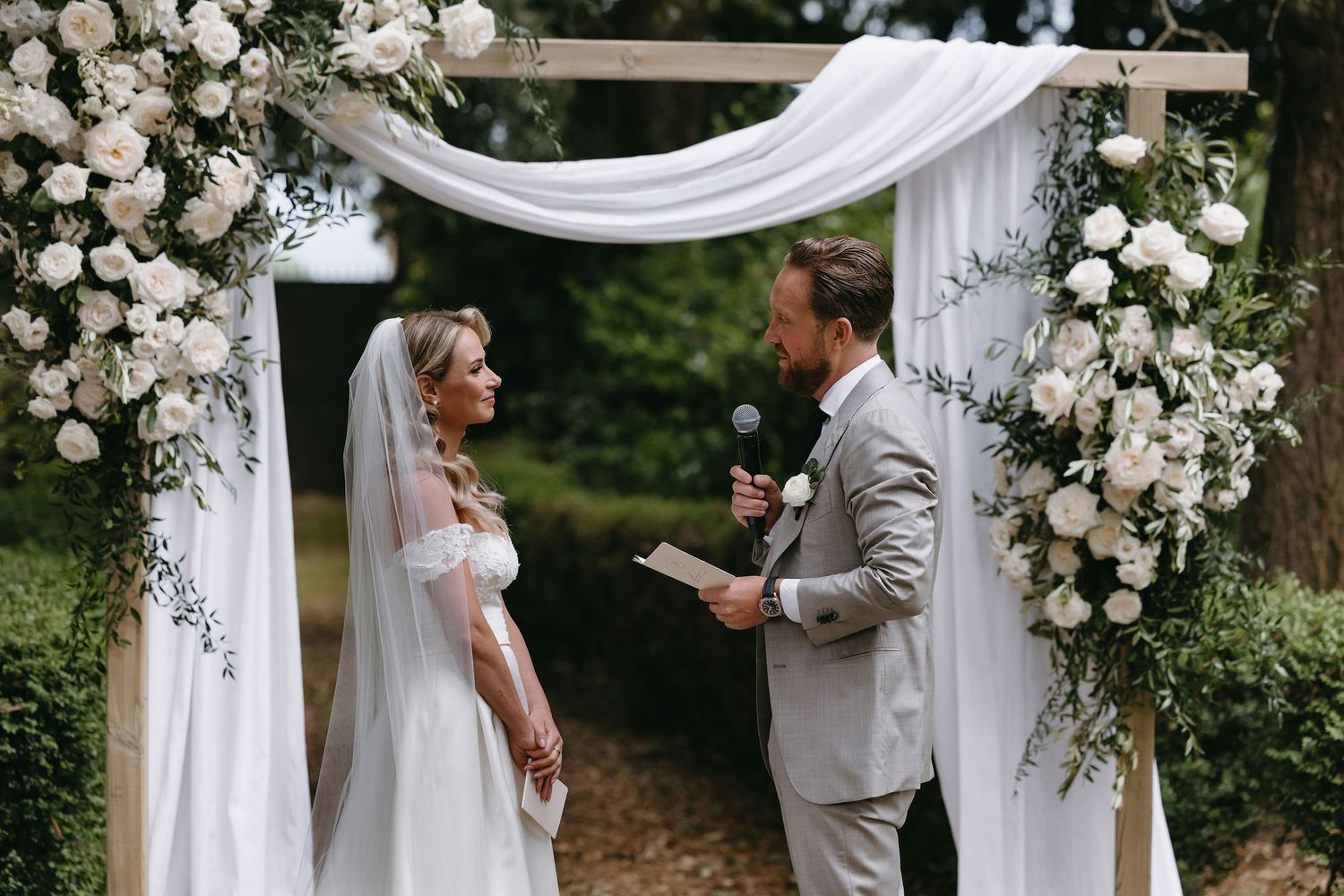 Wedding ceremony under white draped arch decorated with roses as couple exchanges vows.