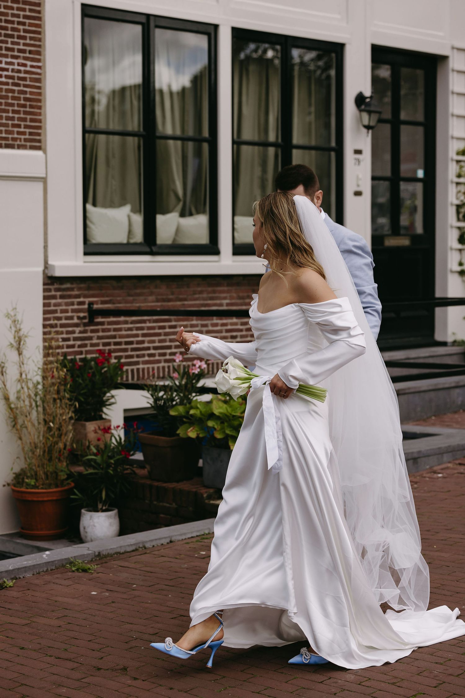 Bride in flowing white dress walking along brick sidewalk with blue heels.