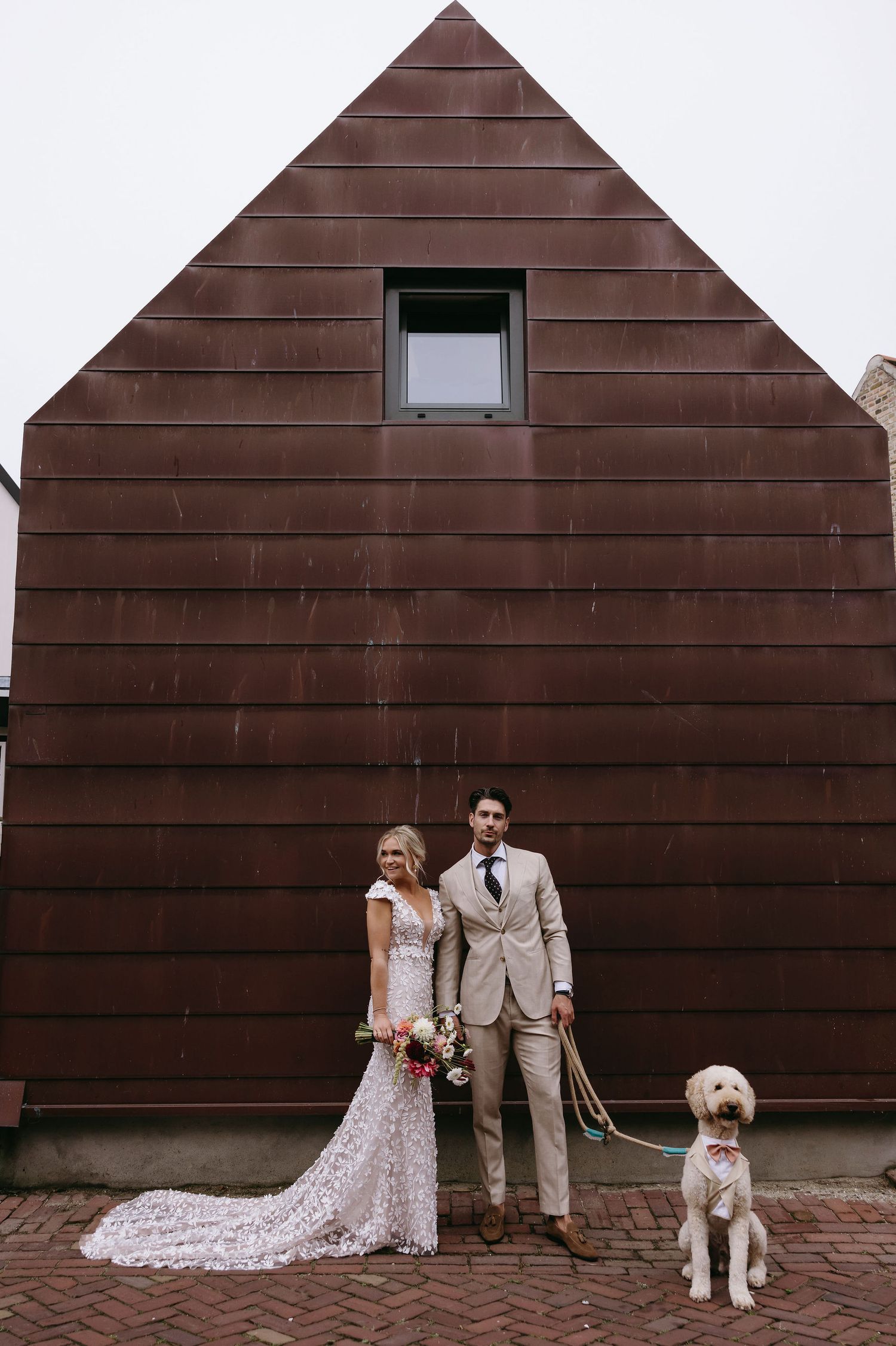 A couple and their white dog pose together in front of a brown wooden wall backdrop.