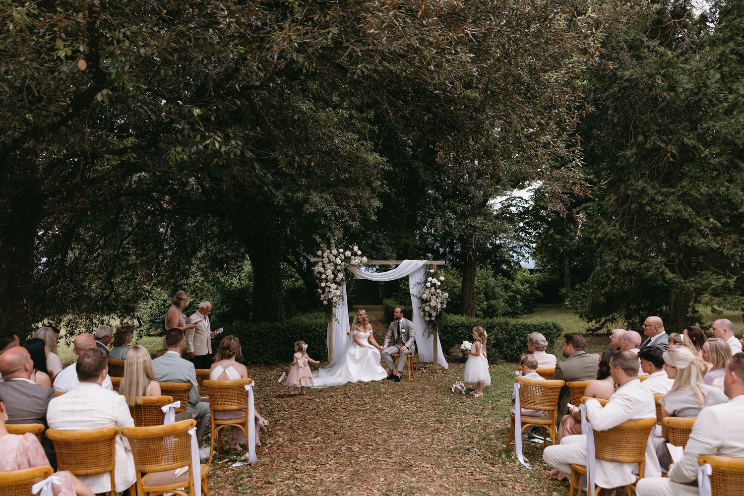 Outdoor wedding ceremony with wooden chairs arranged in rows under mature trees.
