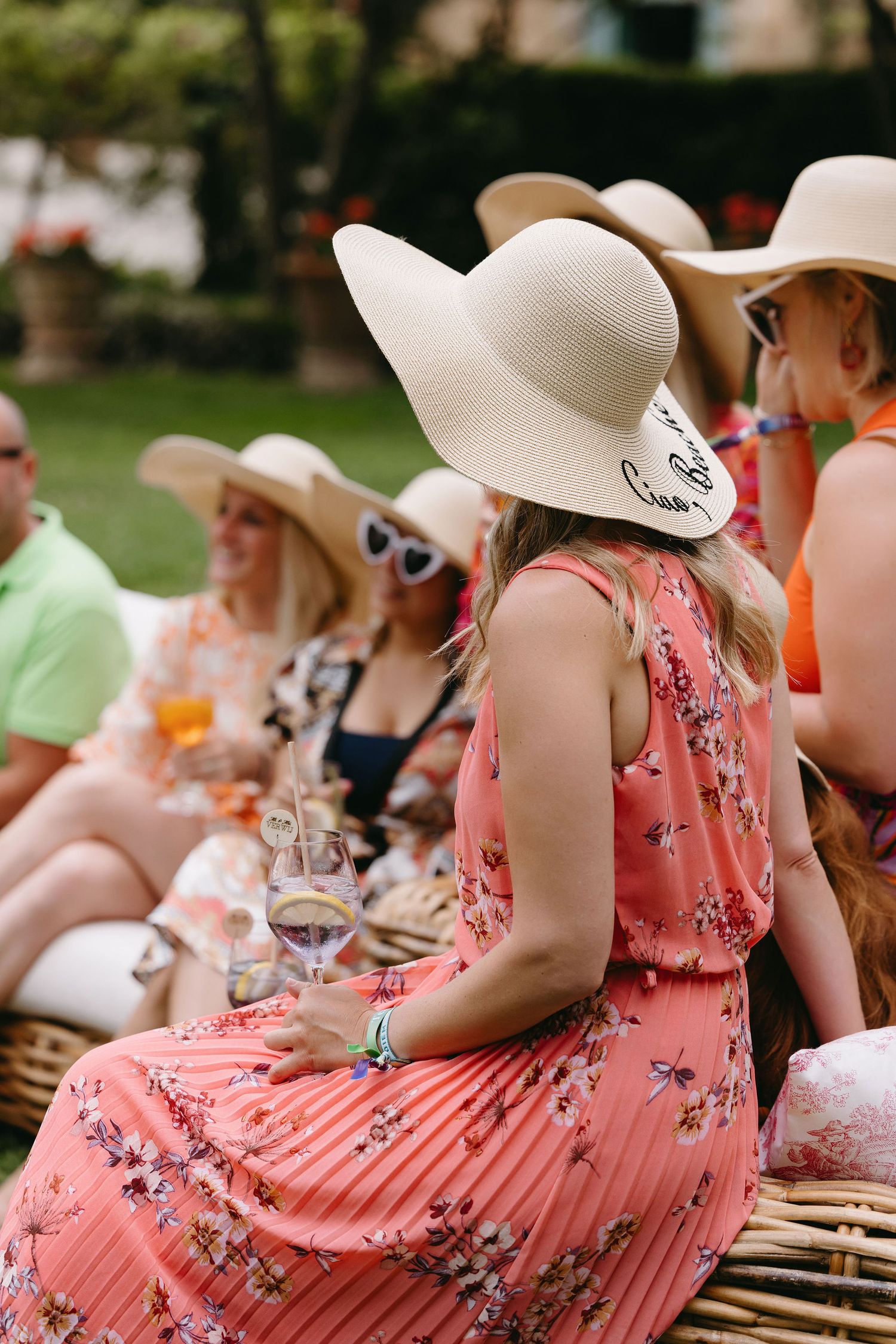 Wedding guests wearing pink floral dresses and wide-brimmed white sun hats seated at an outdoor table.