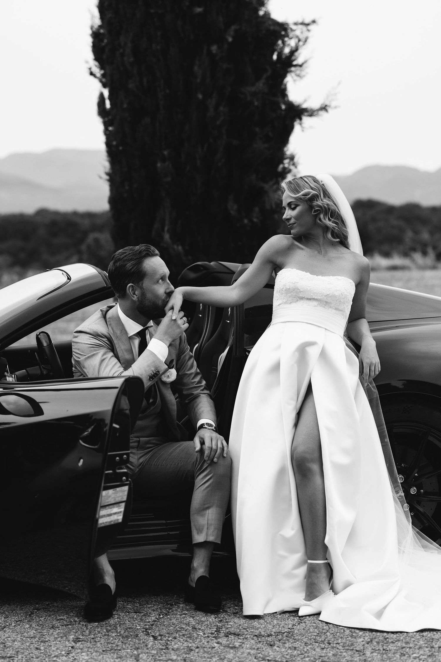 Romantic black and white wedding photo of a couple posing with a convertible car against a mountain backdrop.