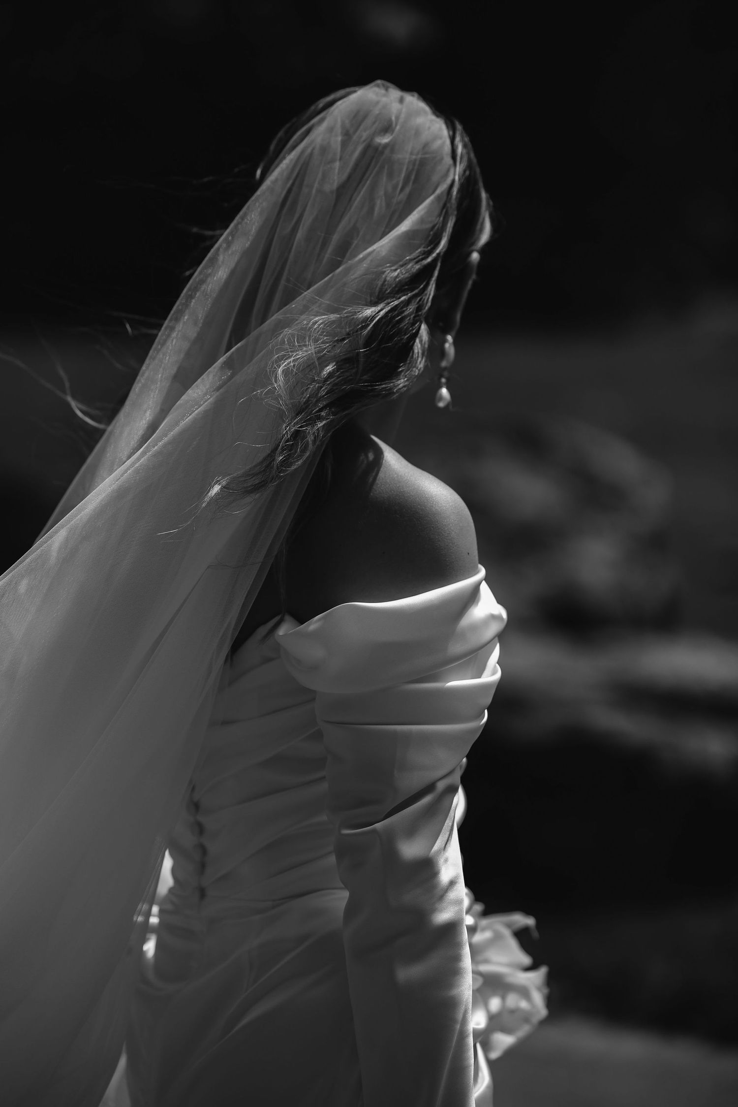 Black and white portrait of a bride in an off-shoulder wedding gown with flowing veil caught in a soft breeze.