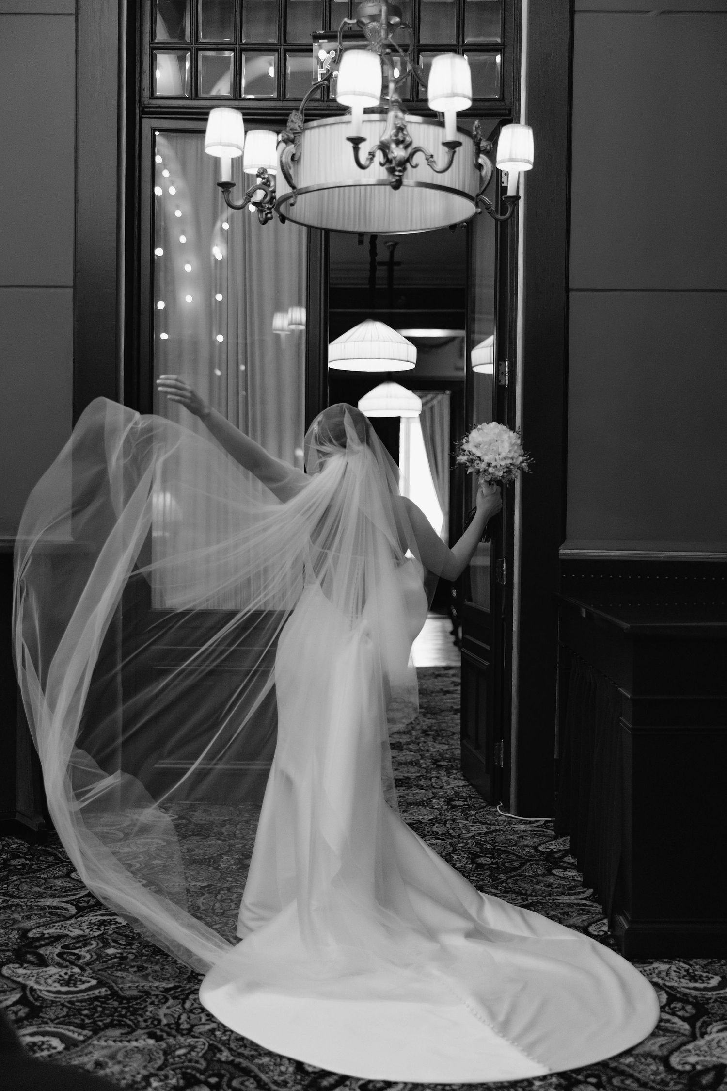 A bride in a flowing white gown with a long veil stands near an elegant chandelier in a black and white photograph.