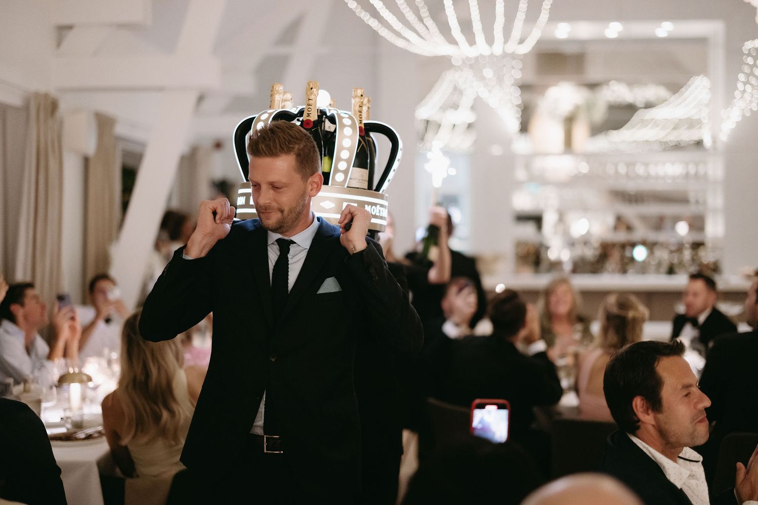 Wedding guests enjoy dinner under crystal chandeliers in an elegant white reception room.