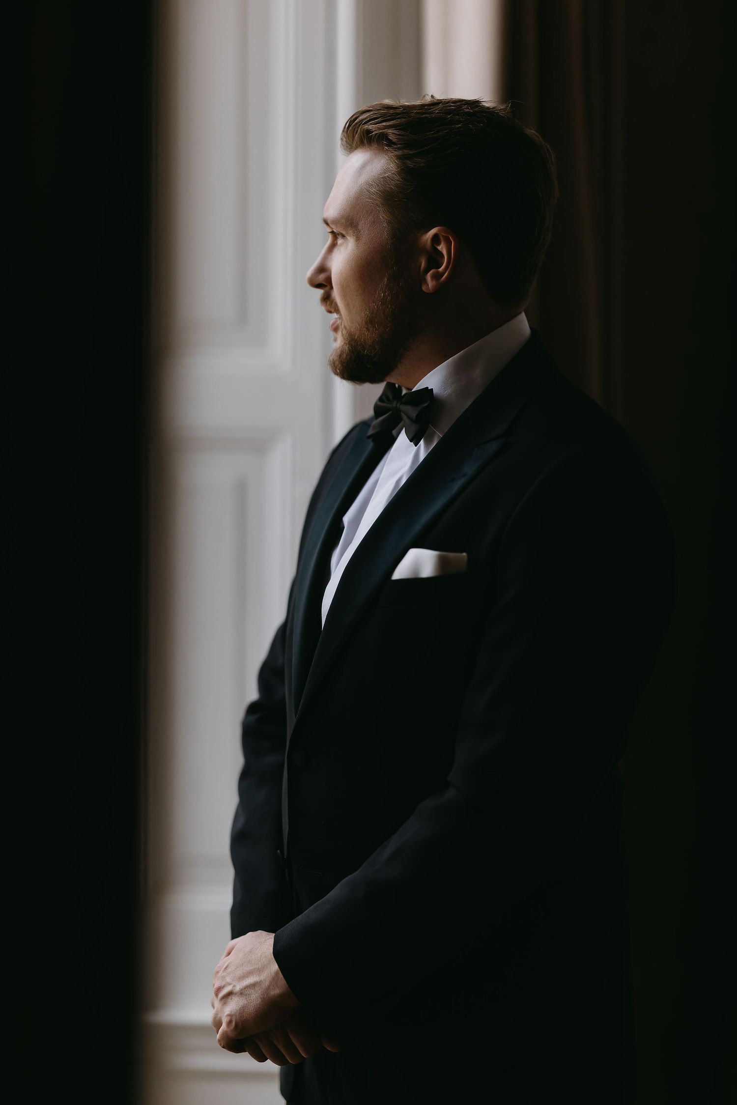Silhouette of a groom in formal attire standing by a window in dramatic lighting.