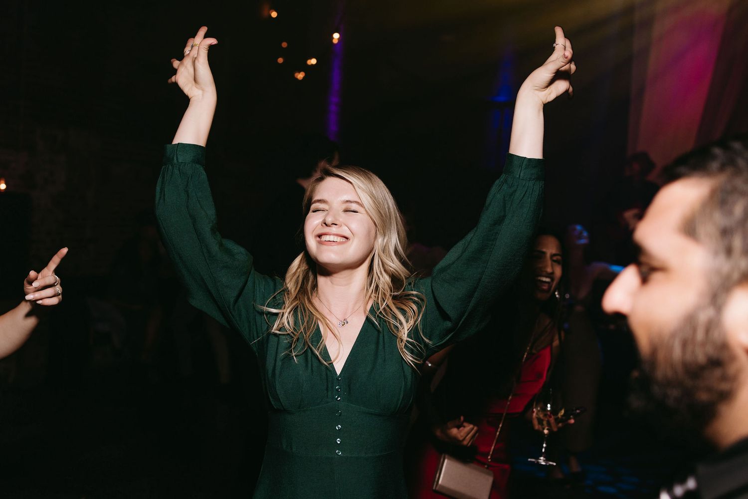A guest joyfully dances at a wedding reception with arms raised in celebration while wearing an emerald green dress.