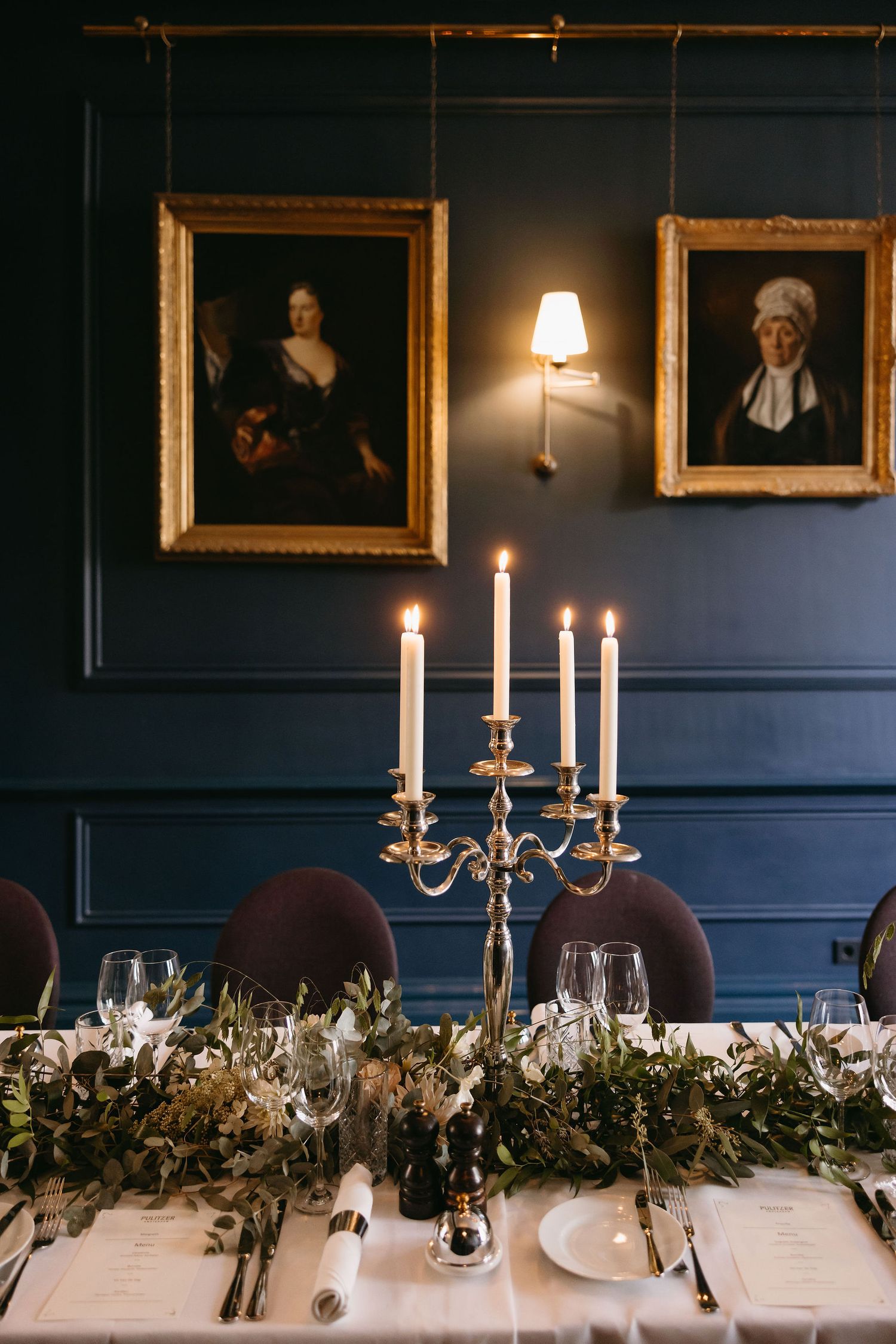 Formal dining table decorated with greenery garland and silver candelabras against navy blue walls.