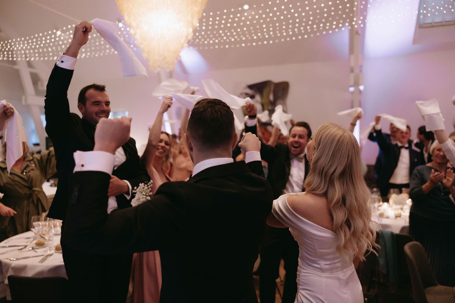 Wedding reception guests celebrate with raised arms under twinkling lights in an indoor venue.