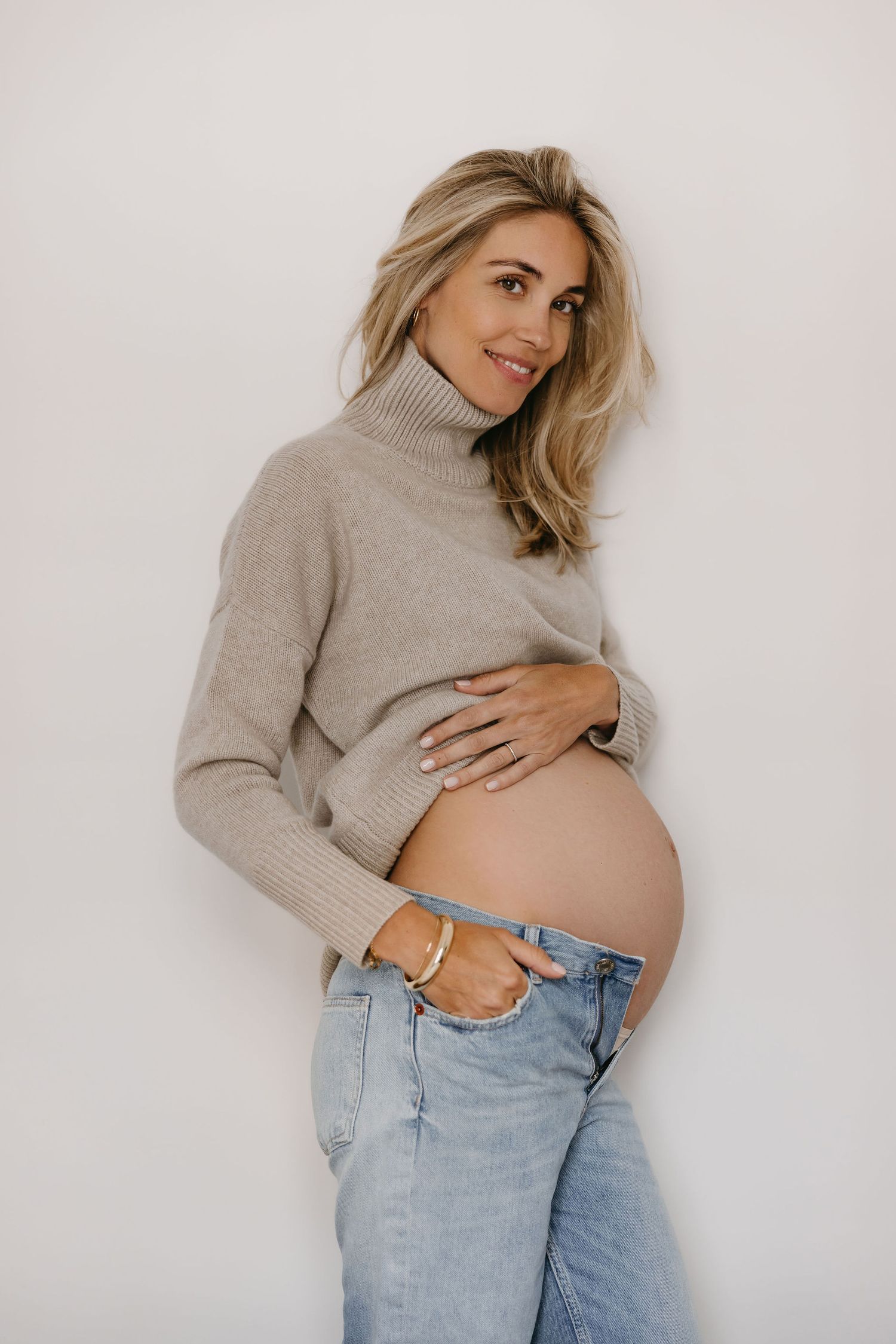 Pregnant woman in beige turtleneck sweater and jeans poses against white wall.