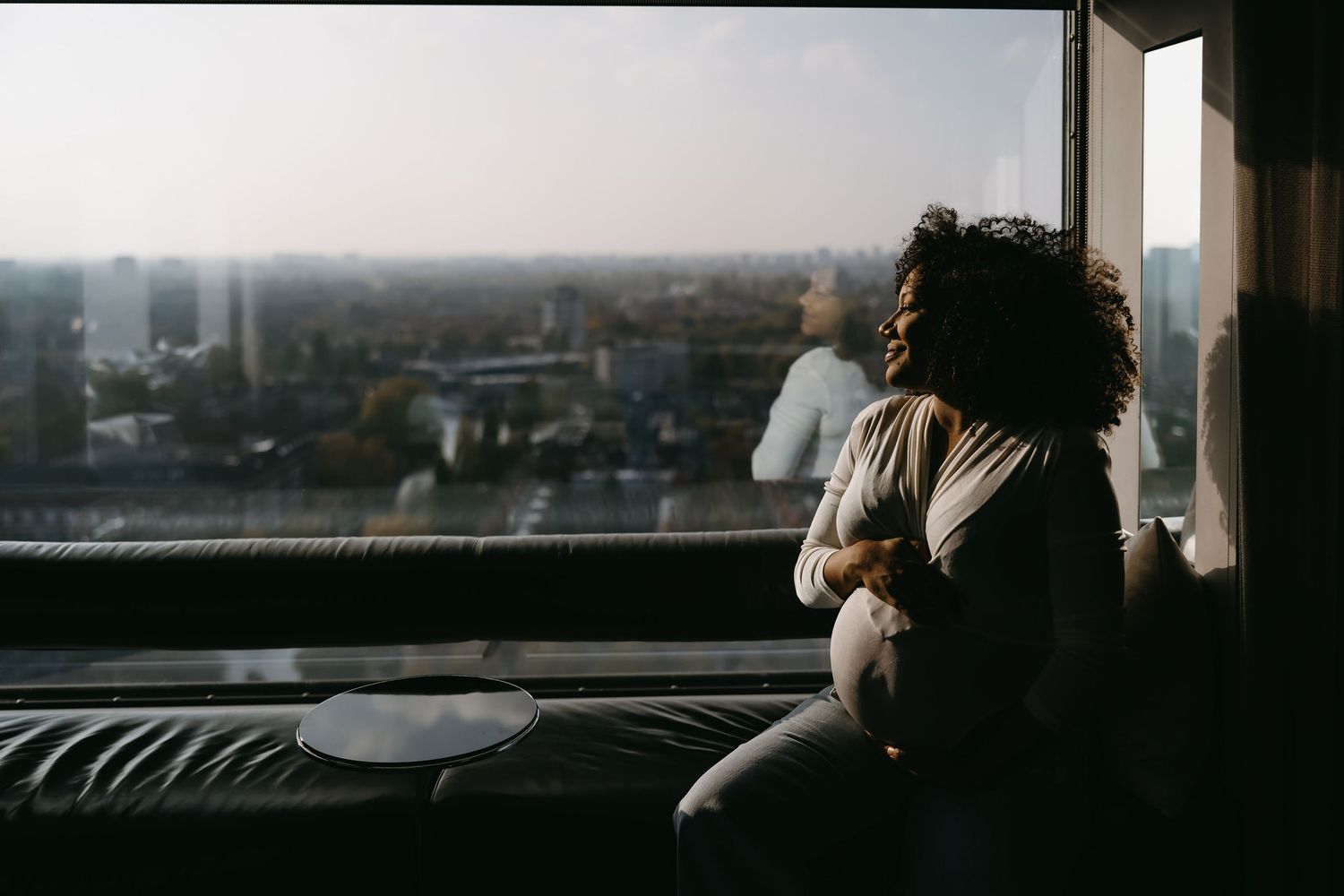 Person gazes out large window at urban cityscape from high vantage point.