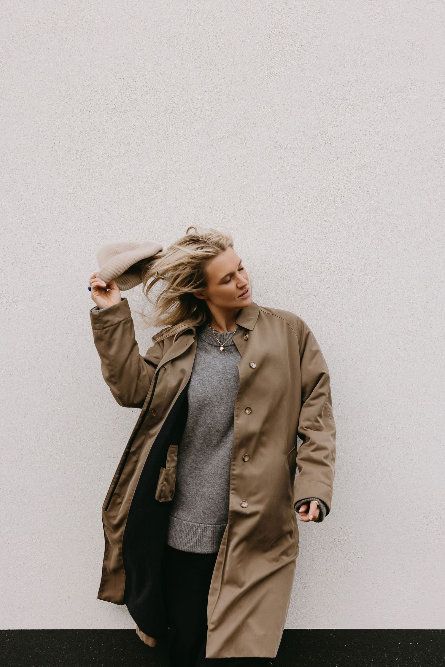 Playful movements in tan coat against white wall in minimalist fashion photoshoot.