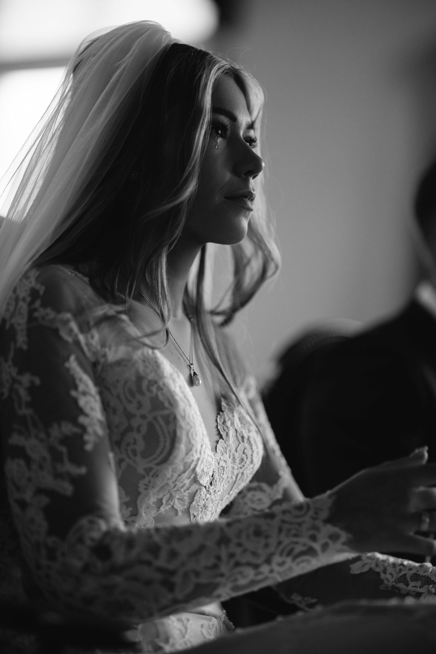 A bride in a white lace gown poses in dramatic window light during bridal preparations.