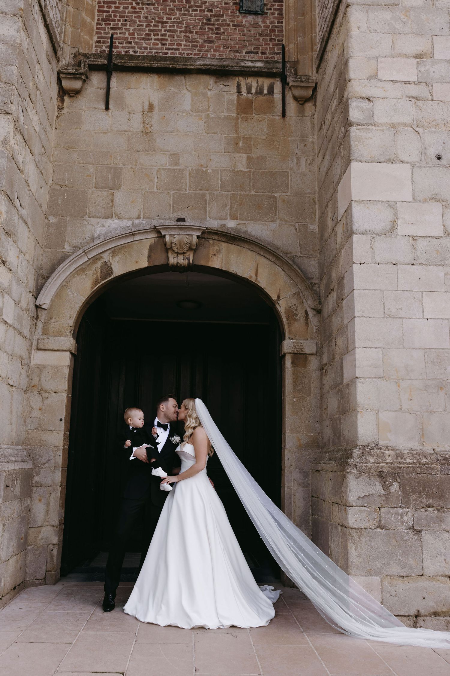 A wedding couple stands in an elegant stone archway entrance of a historic building with a long flowing veil.