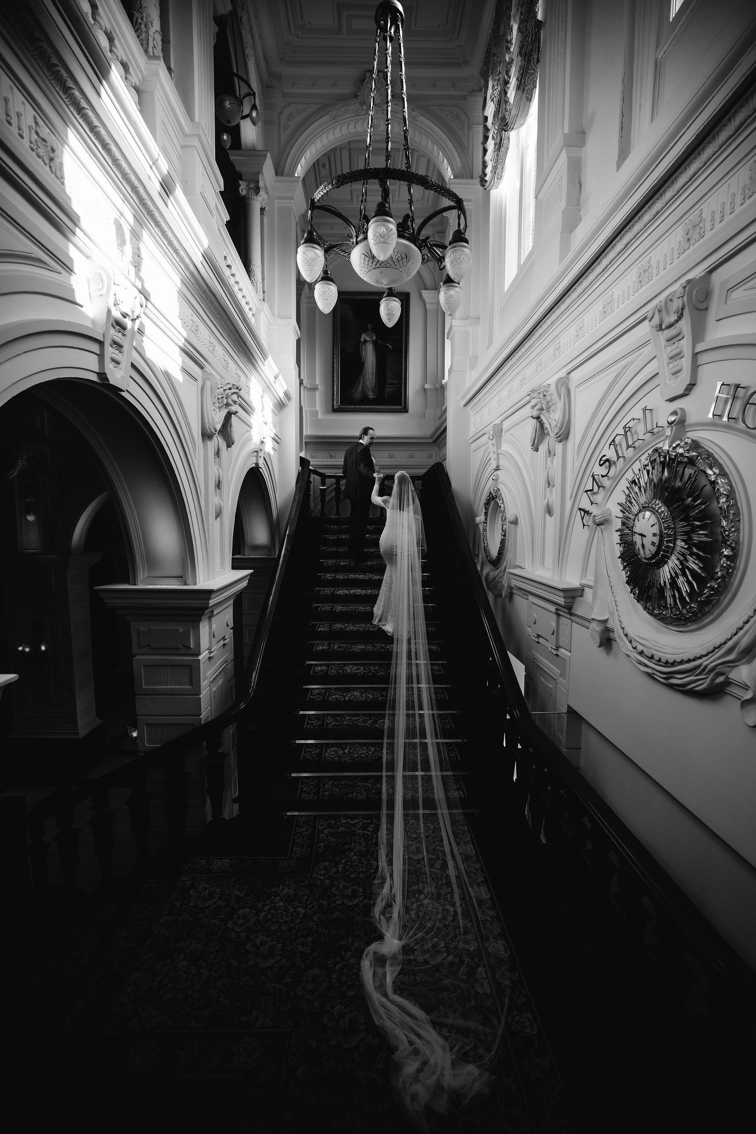 Black and white photo of an ornate hallway with dramatic staircase and hanging chandelier with a trailing bridal veil.