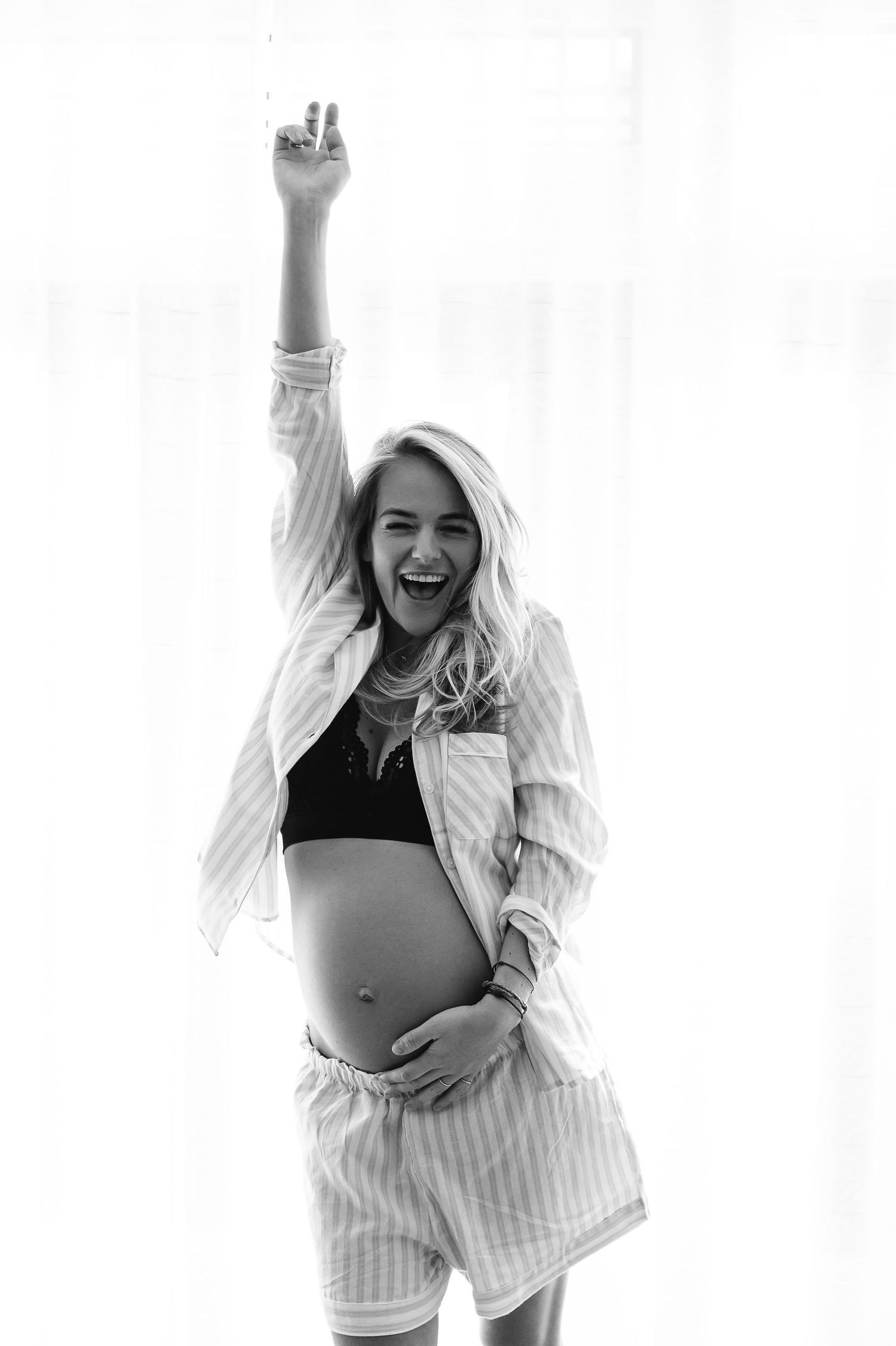 Joyful expecting mother in striped pajamas and white jacket raises arm in celebration against white backdrop.