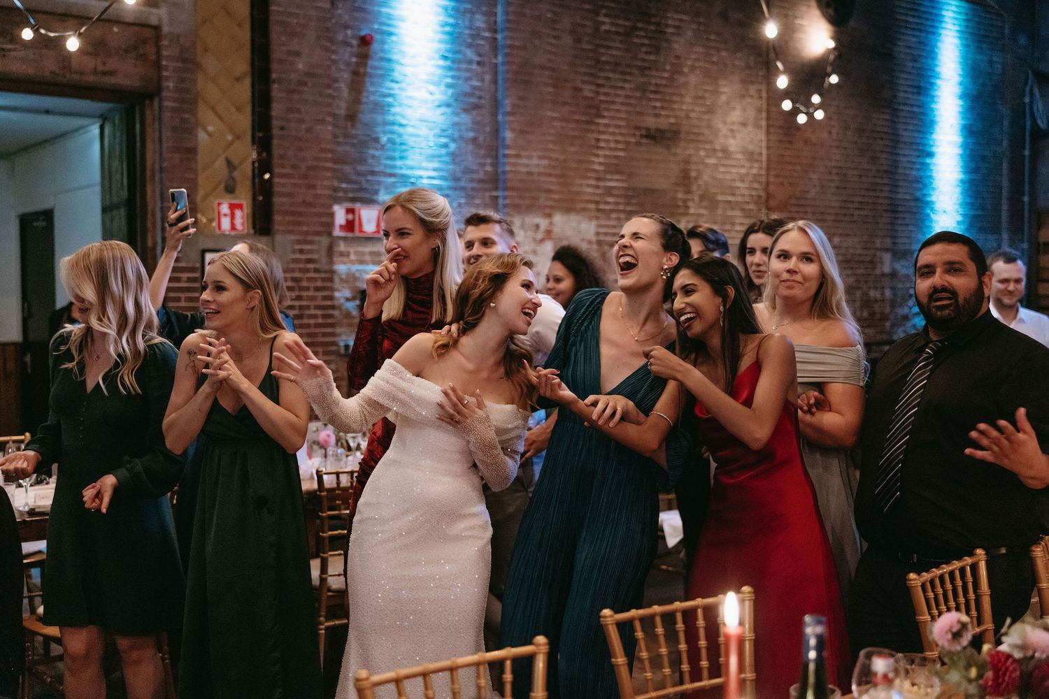 Wedding guests gather in a rustic brick venue with blue lighting, reacting joyfully during a celebration.