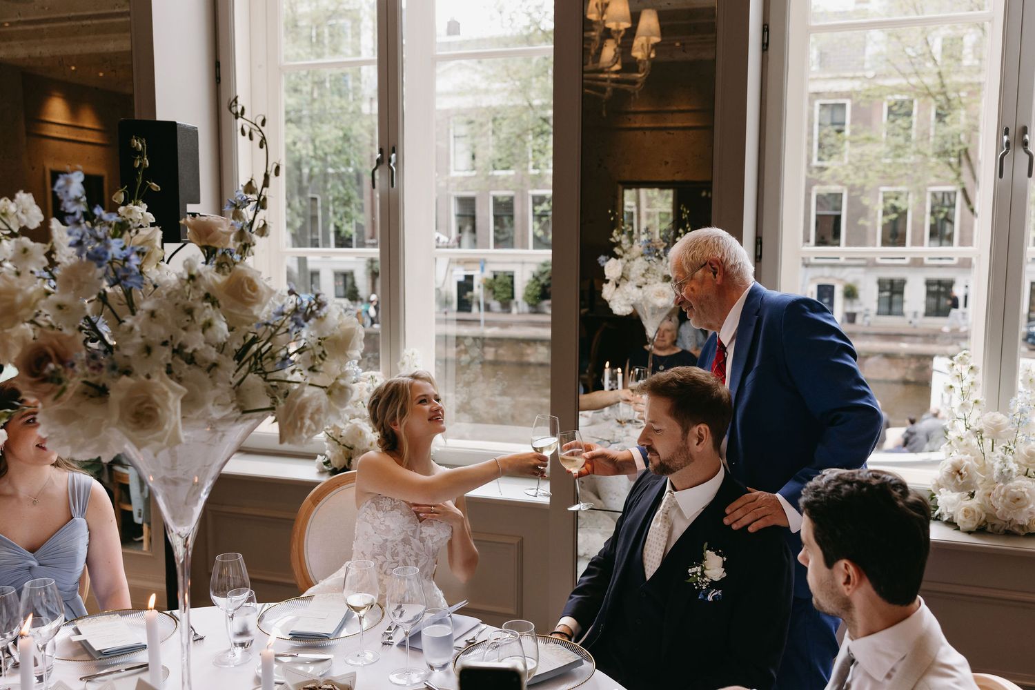 Wedding guests enjoying a reception dinner in an elegant venue with large windows and floral arrangements.